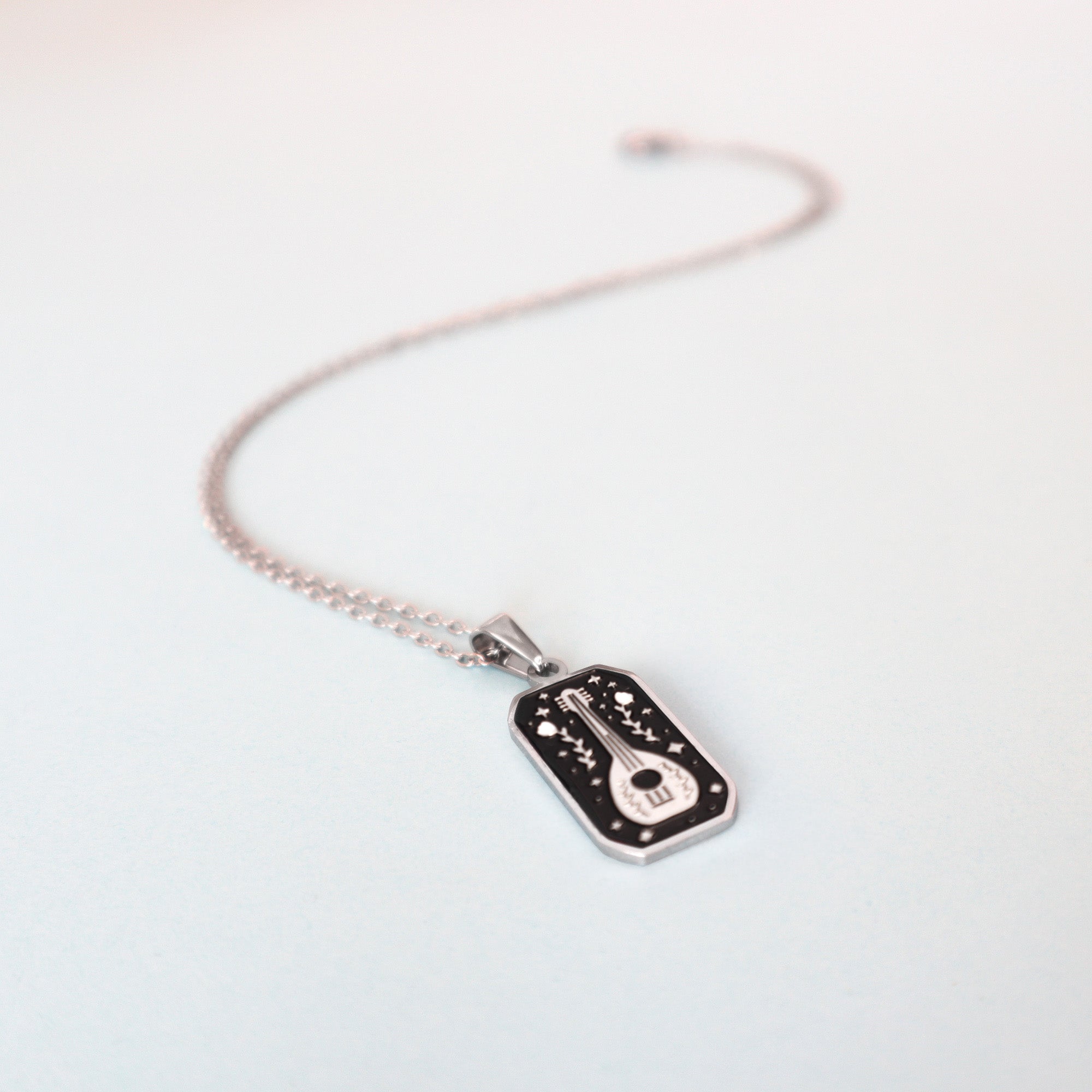 The Bard Pendant Enamel Necklace by Papio Press features a rectangular black and silver pendant with a white guitar and star designs, perfect for fantasy lovers. Displayed on a light pastel background.