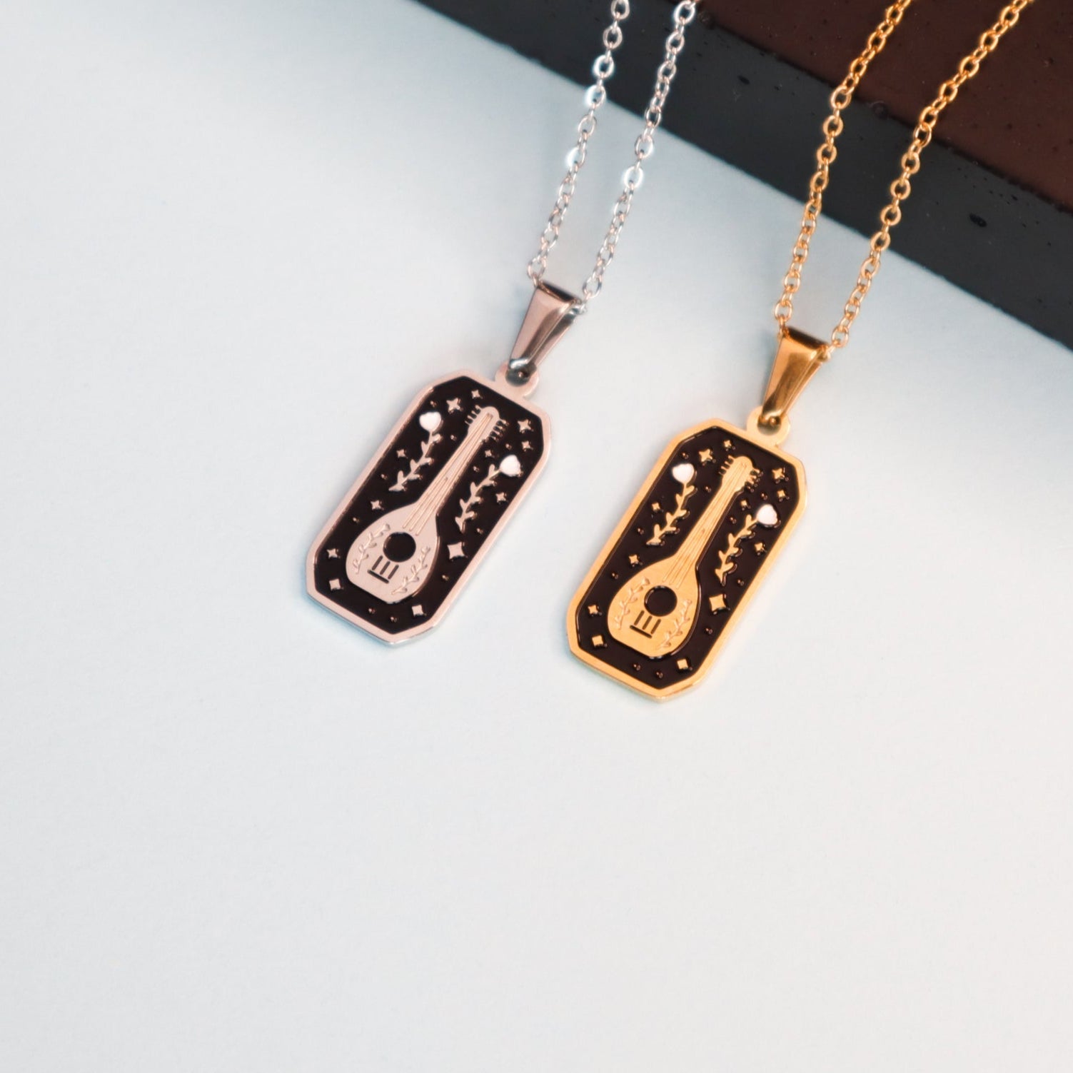 The Bard Pendant Enamel Necklaces by Papio Press—two necklaces with engraved guitar and star designs, in 18K gold-plated or silver finishes—make perfect fantasy game accessories, shown on a light surface near a dark brown object.