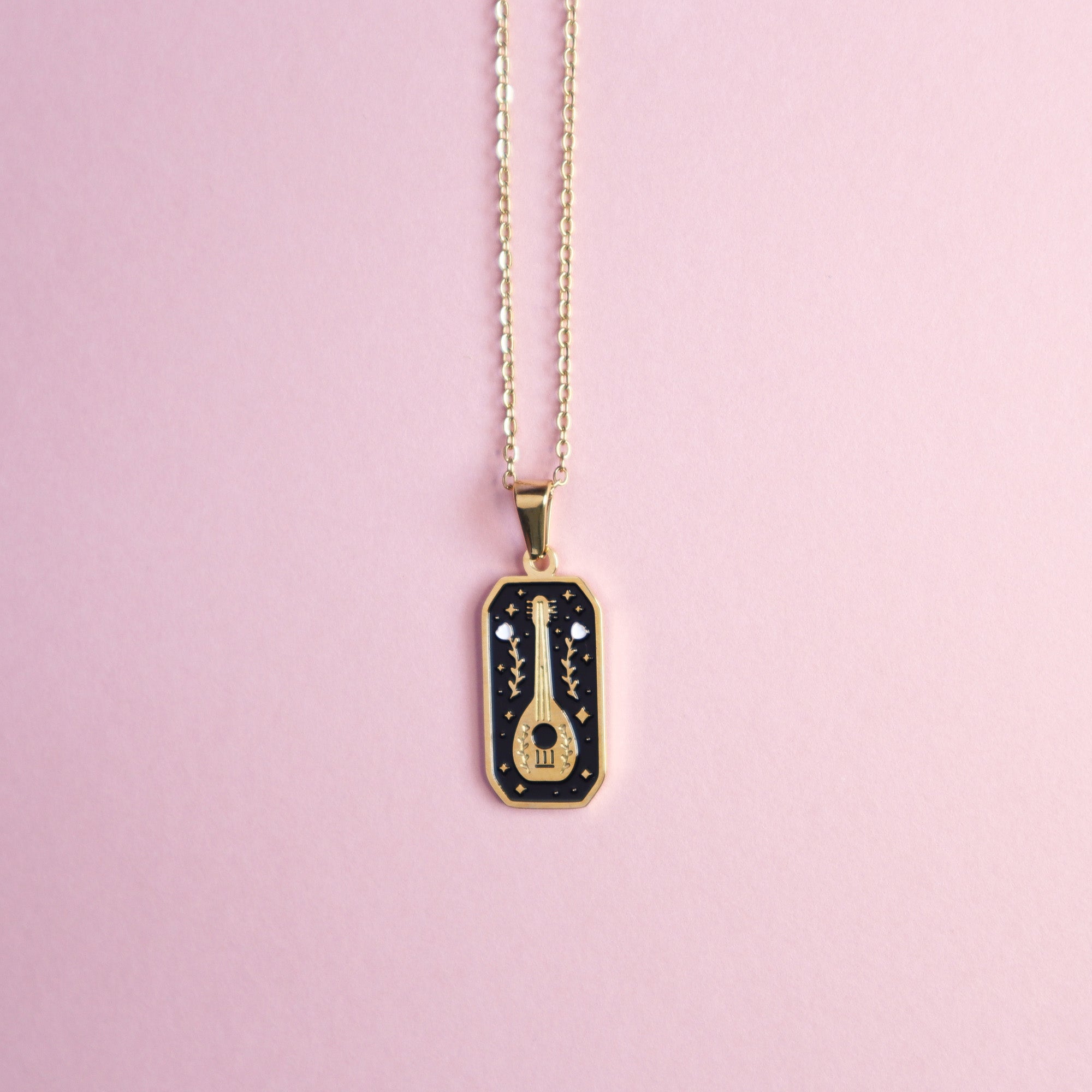 The Bard Pendant Enamel Necklace by Papio Press features a rectangular, 18K gold plated pendant with a black background, tiny stars, and a gold guitar—ideal for fantasy game fans. Shown on a light pink surface.