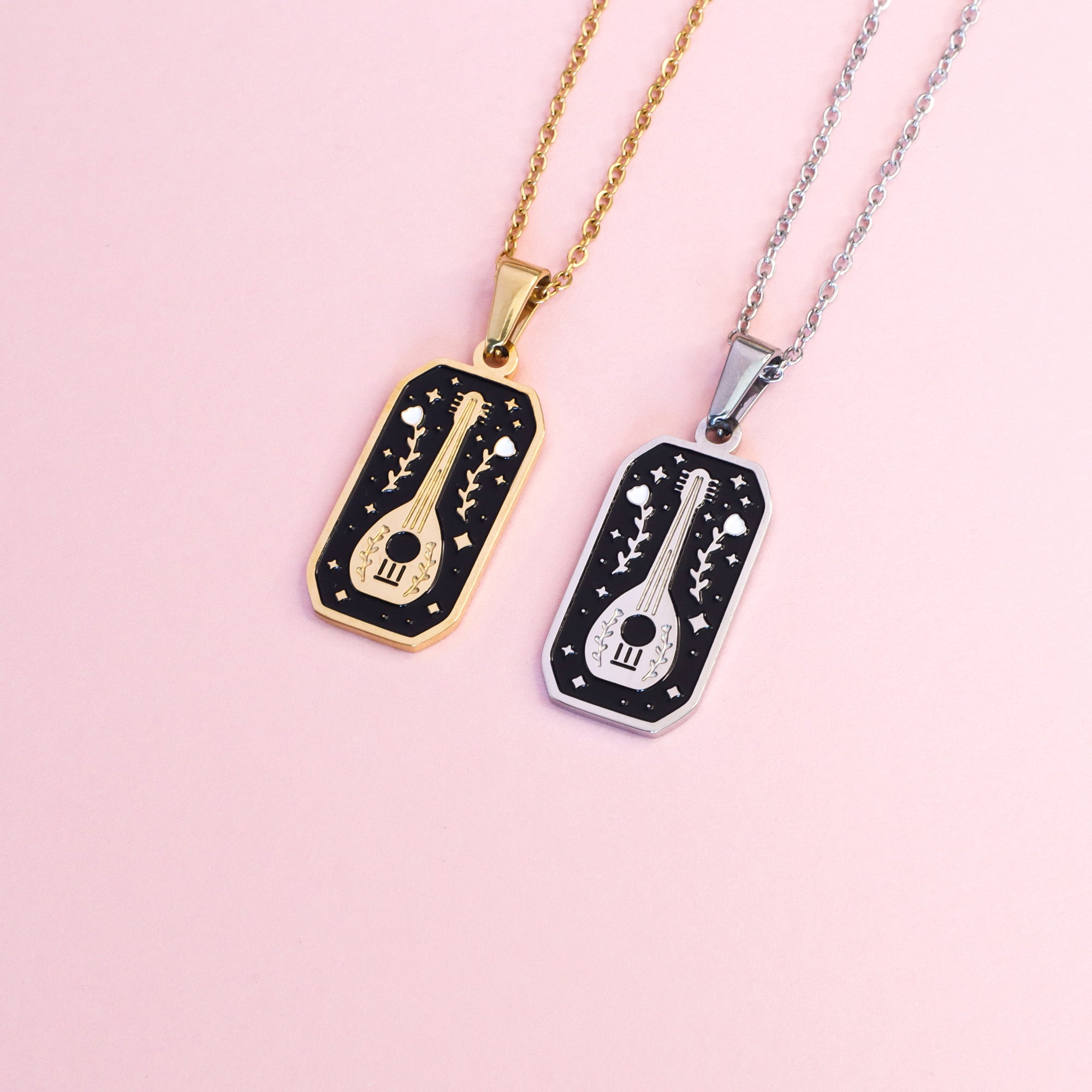 The Bard Pendant Enamel Necklaces by Papio Press—two necklaces with engraved guitar and star designs, in 18K gold-plated or silver finishes—make perfect fantasy game accessories, shown on a light pink surface.