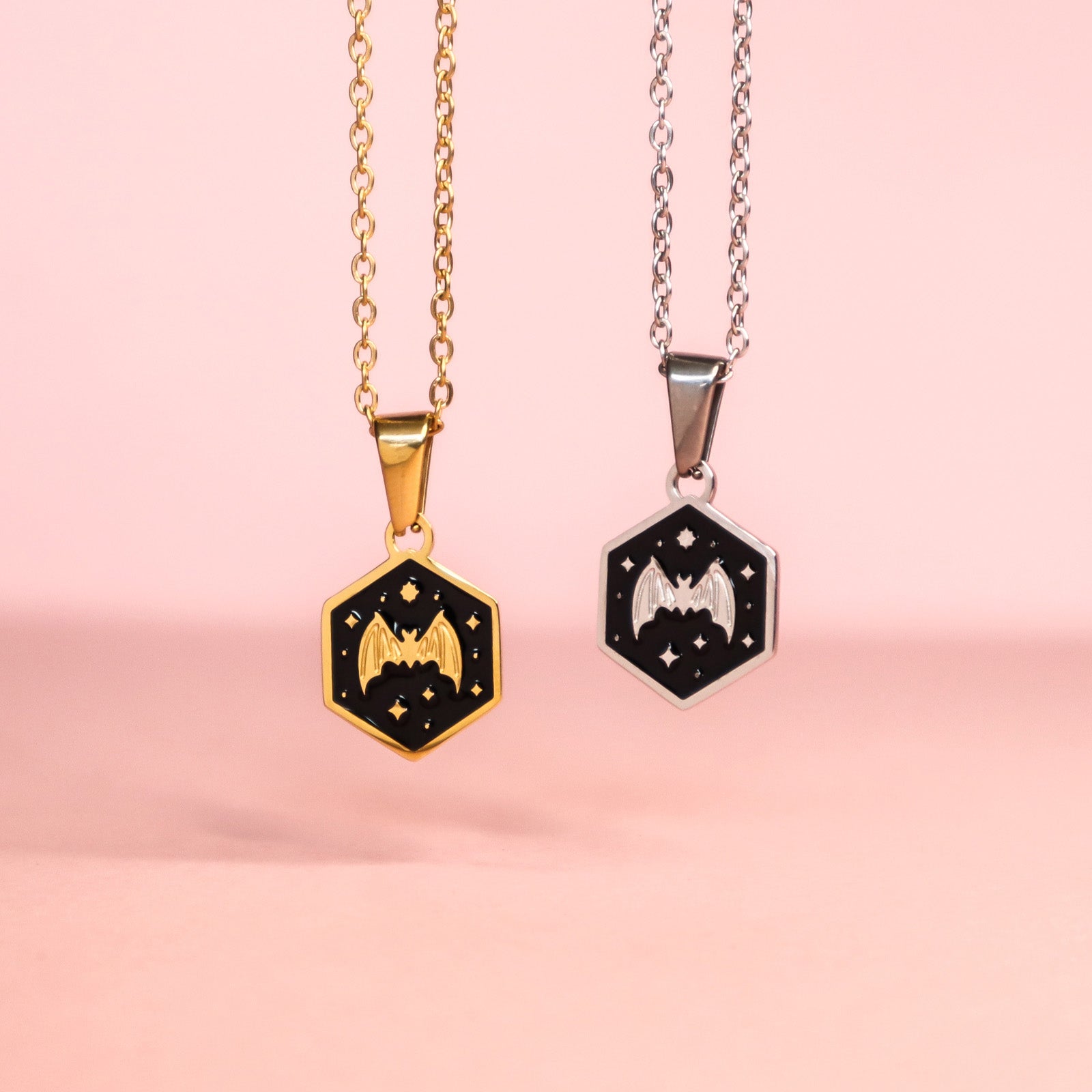 Two Papio Press Mini Hexagon Bat Pendants, one gold and one silver, each with an enamel bat and stars on a black background, displayed on chains against a soft pink surface.