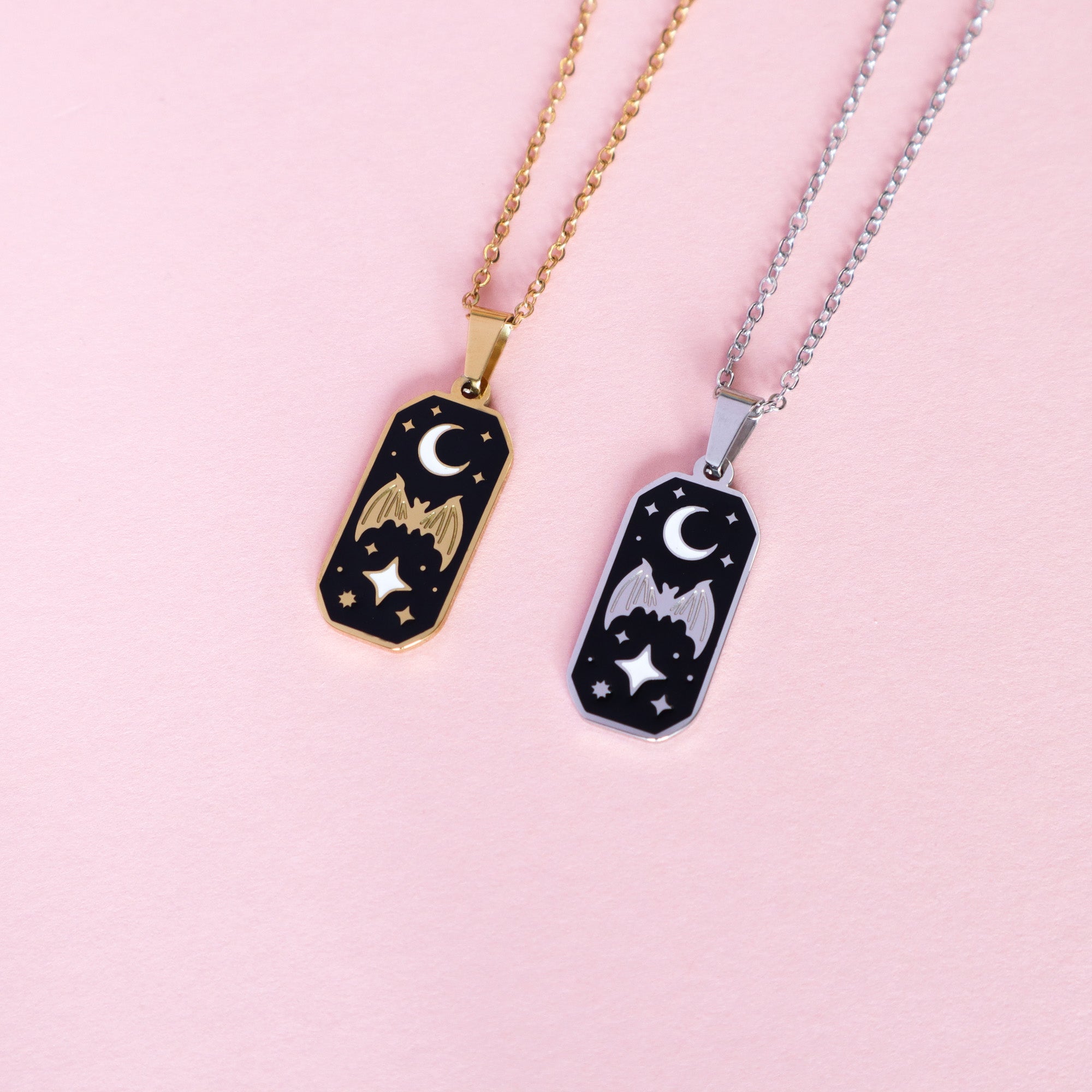 Papio Presss Bat & Moon Enamel Necklace features gold and silver rectangular pendants with a bat, stars, and crescent moon on black enamel. Made of stainless steel chains, theyre displayed on a light pink surface.