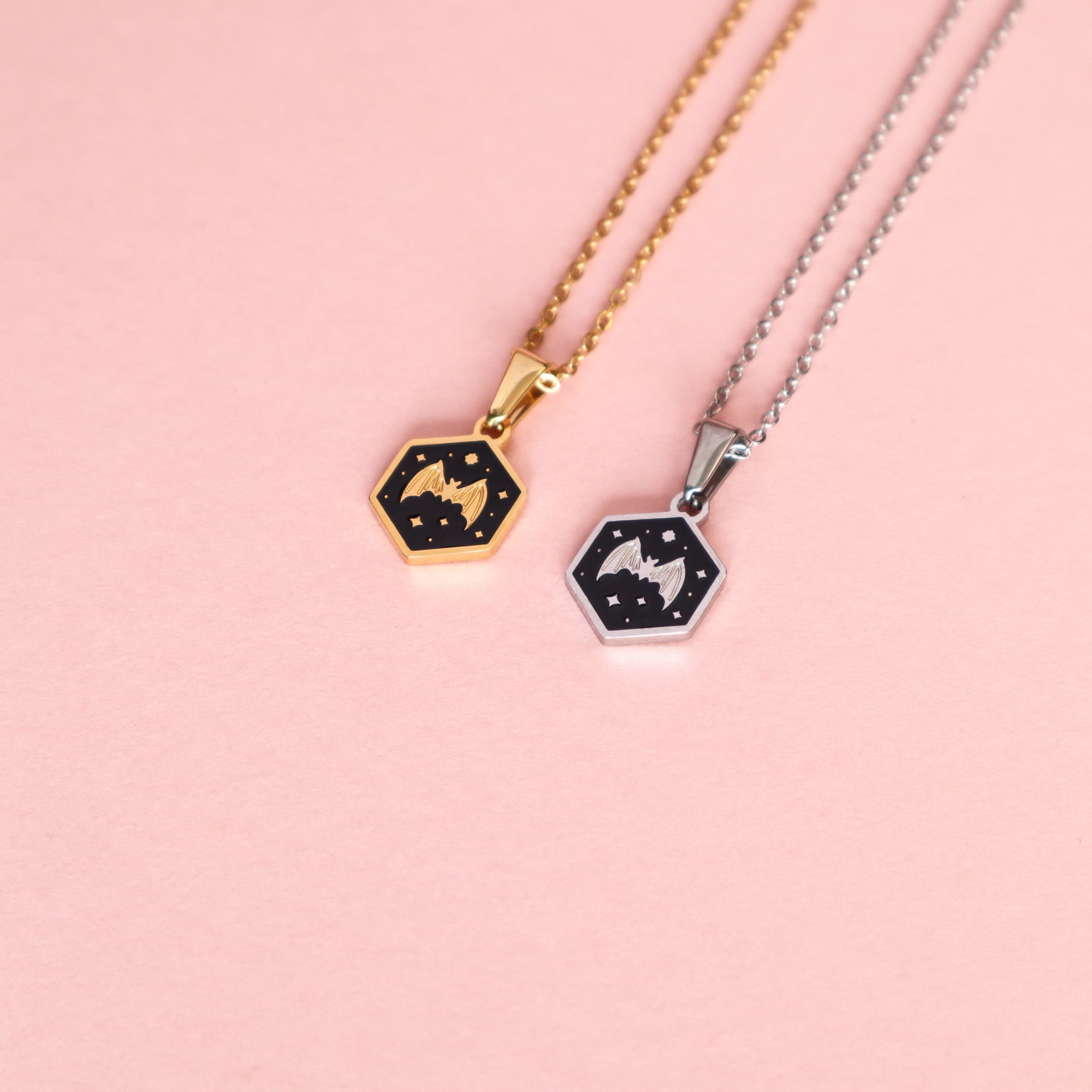 Two Papio Press Mini Hexagon Bat Pendants on a pink background: one with a stainless steel silver chain, one with a gold chain. Both feature a celestial bat design on black, accented with shimmering stars.