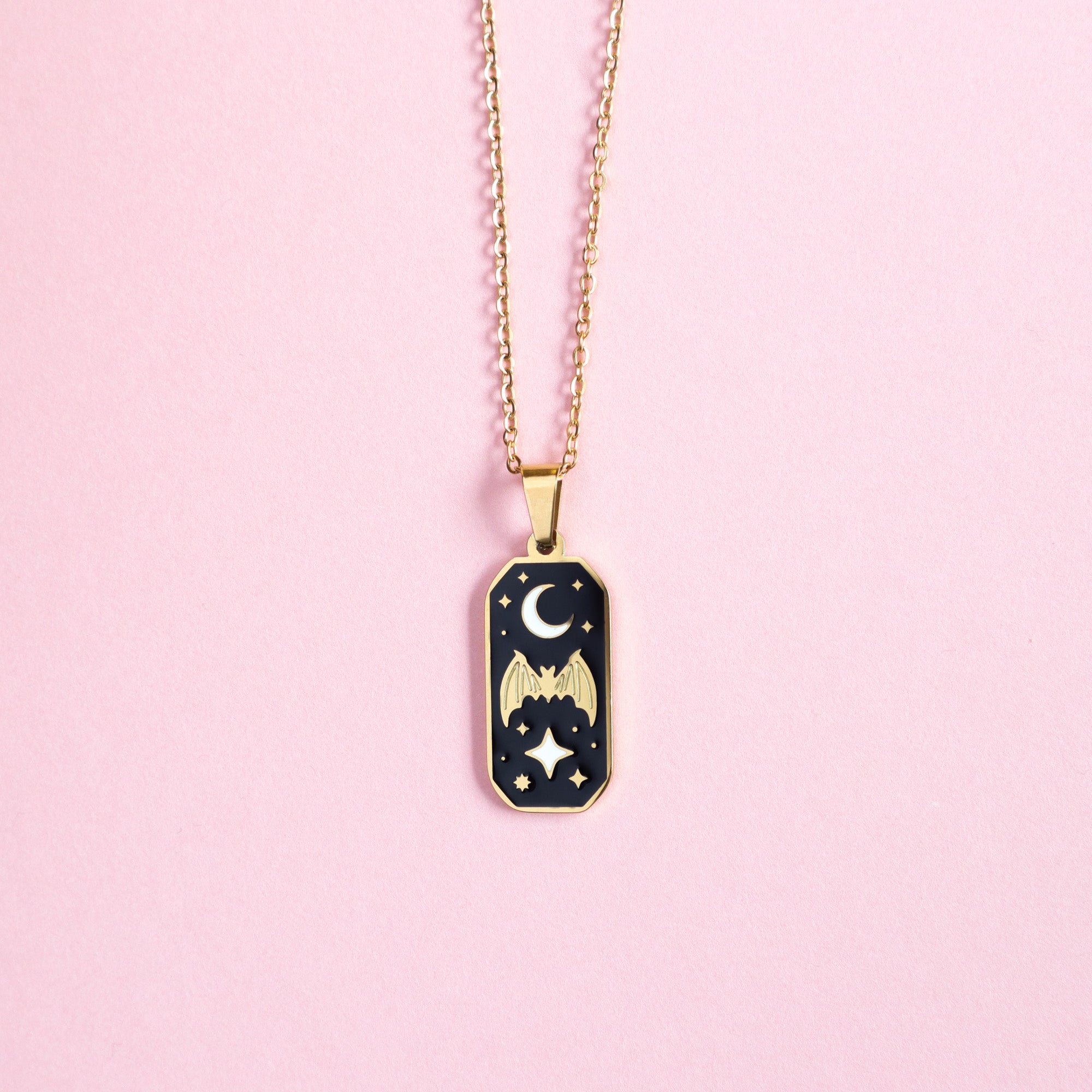 The Bat & Moon Enamel Necklace by Papio Press features a gold chain and black rectangular charm with gold bat wings, crescent moon, and star illustrations, shown on a pink background.
