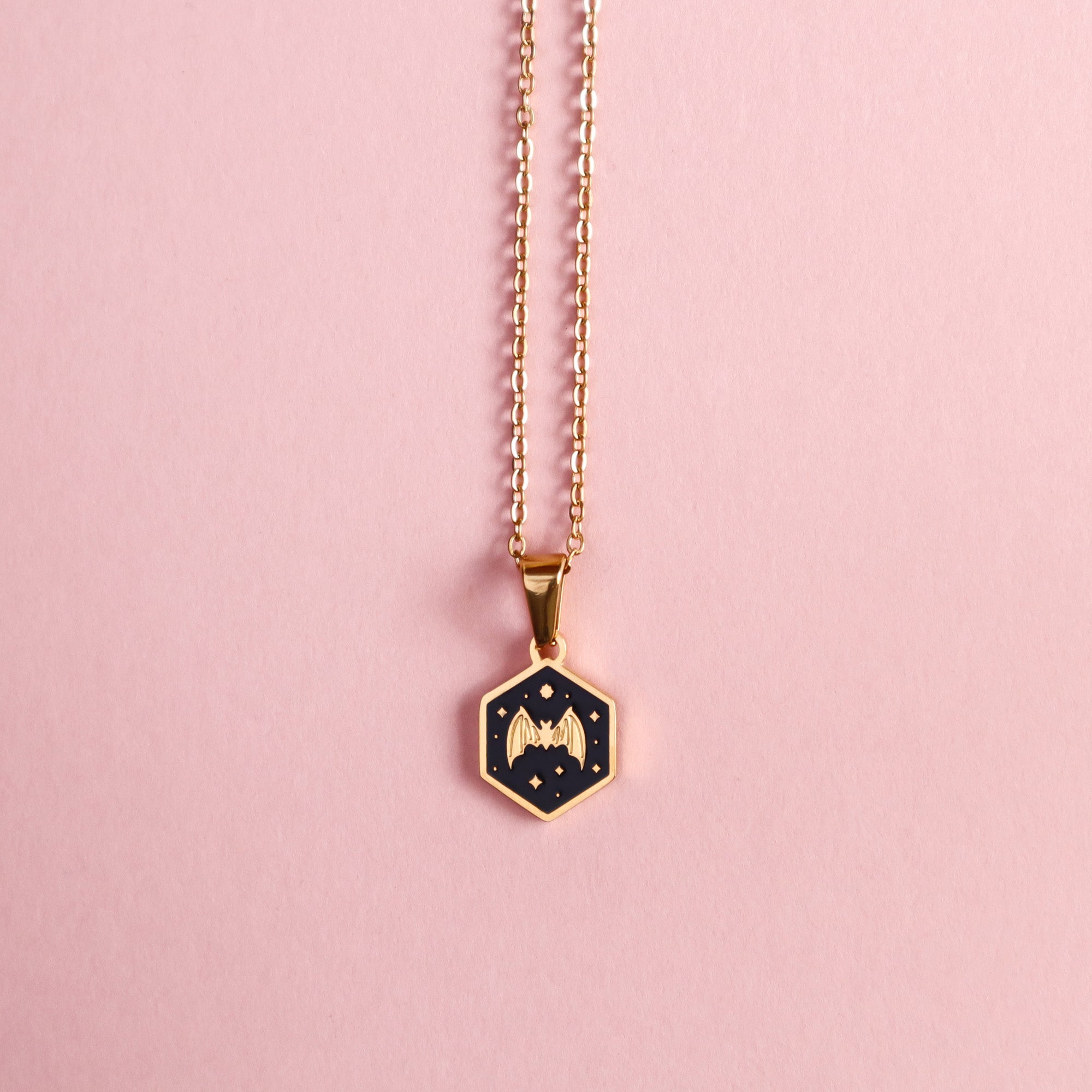 The Mini Hexagon Bat Pendant by Papio Press is a gold enamel necklace featuring a black center, gold bat, and small stars. Displayed on a light pink background, this unique piece makes a striking statement.
