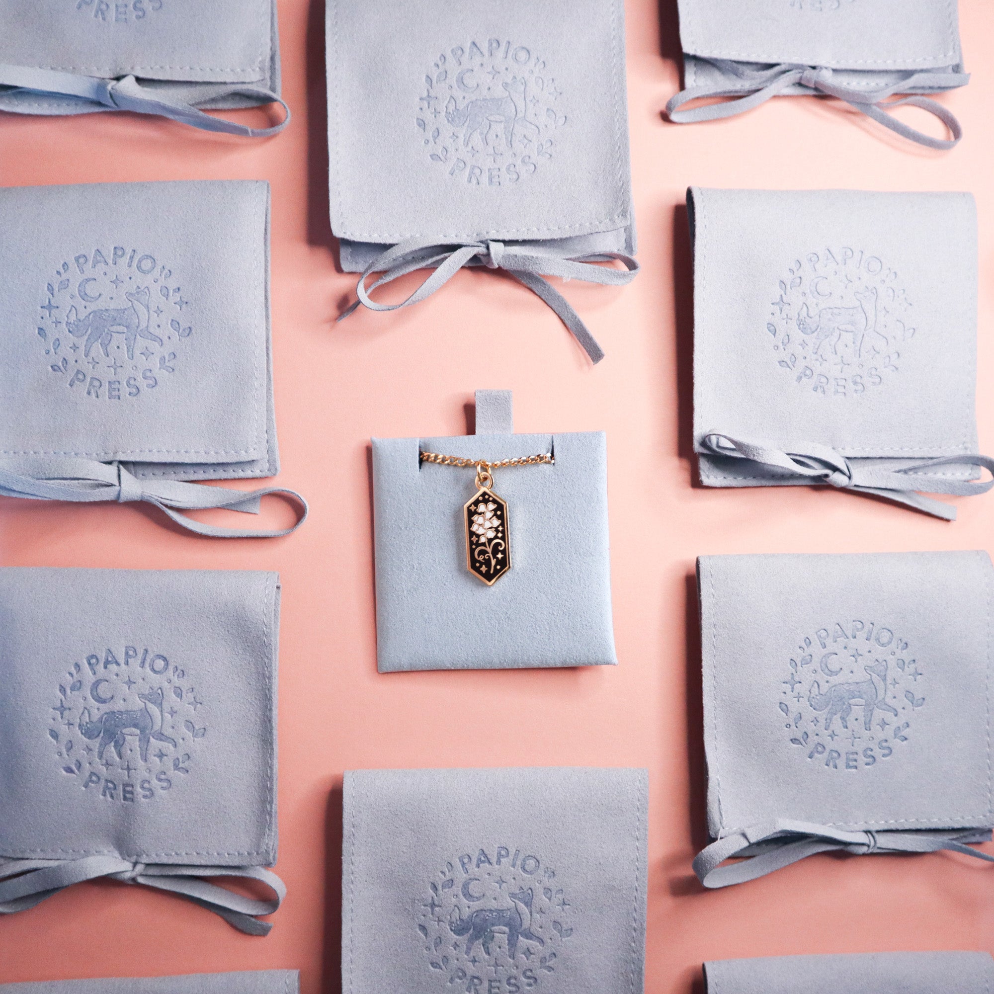 The Lily of the Valley Pendant Enamel Necklace by Papio Press is shown on a light blue pouch, surrounded by similar tied pouches with Papio Press and animal designs, all arranged on a pink background.