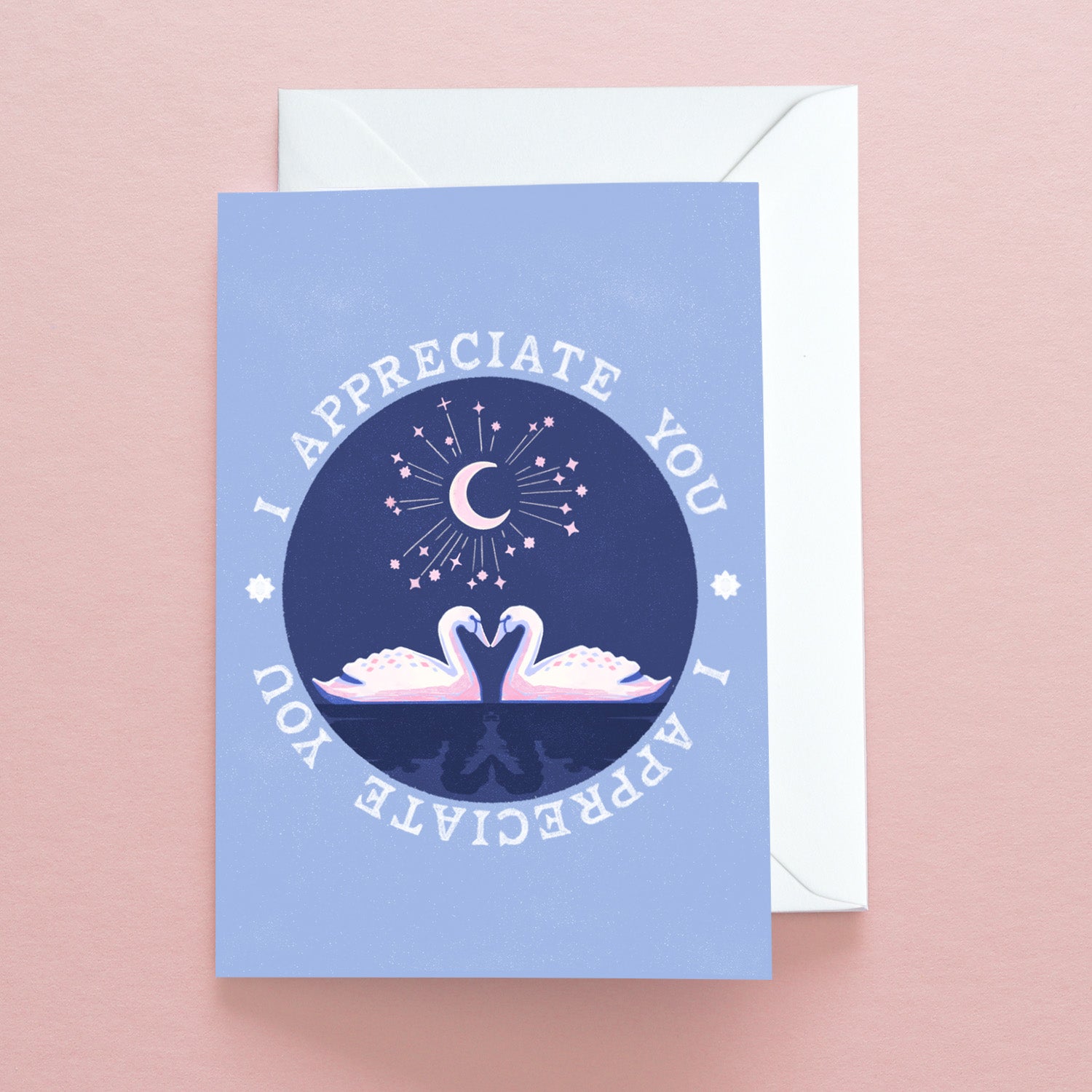The I Appreciate You Swans Greetings Card by Papio Press features two white swans under a starry sky with “I APPRECIATE YOU” above, printed on recycled stock and shown resting on a pink surface.