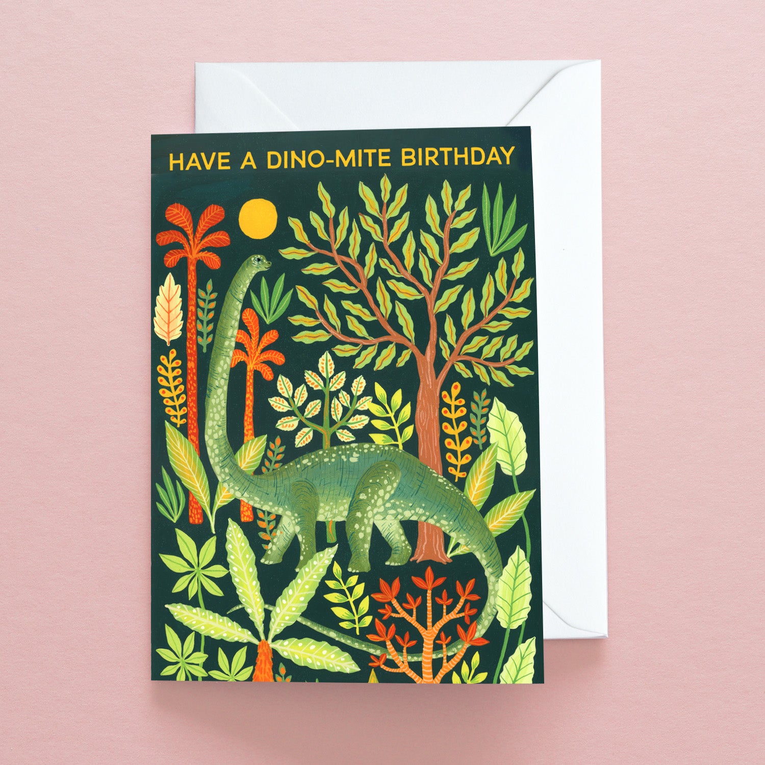 The Have a Dino-Mite Birthday Greetings Card by Papio Press features a green dinosaur among colorful plants on a dark background, with the message Have a Dino-mite Birthday. Printed on recycled stock, it includes an envelope.