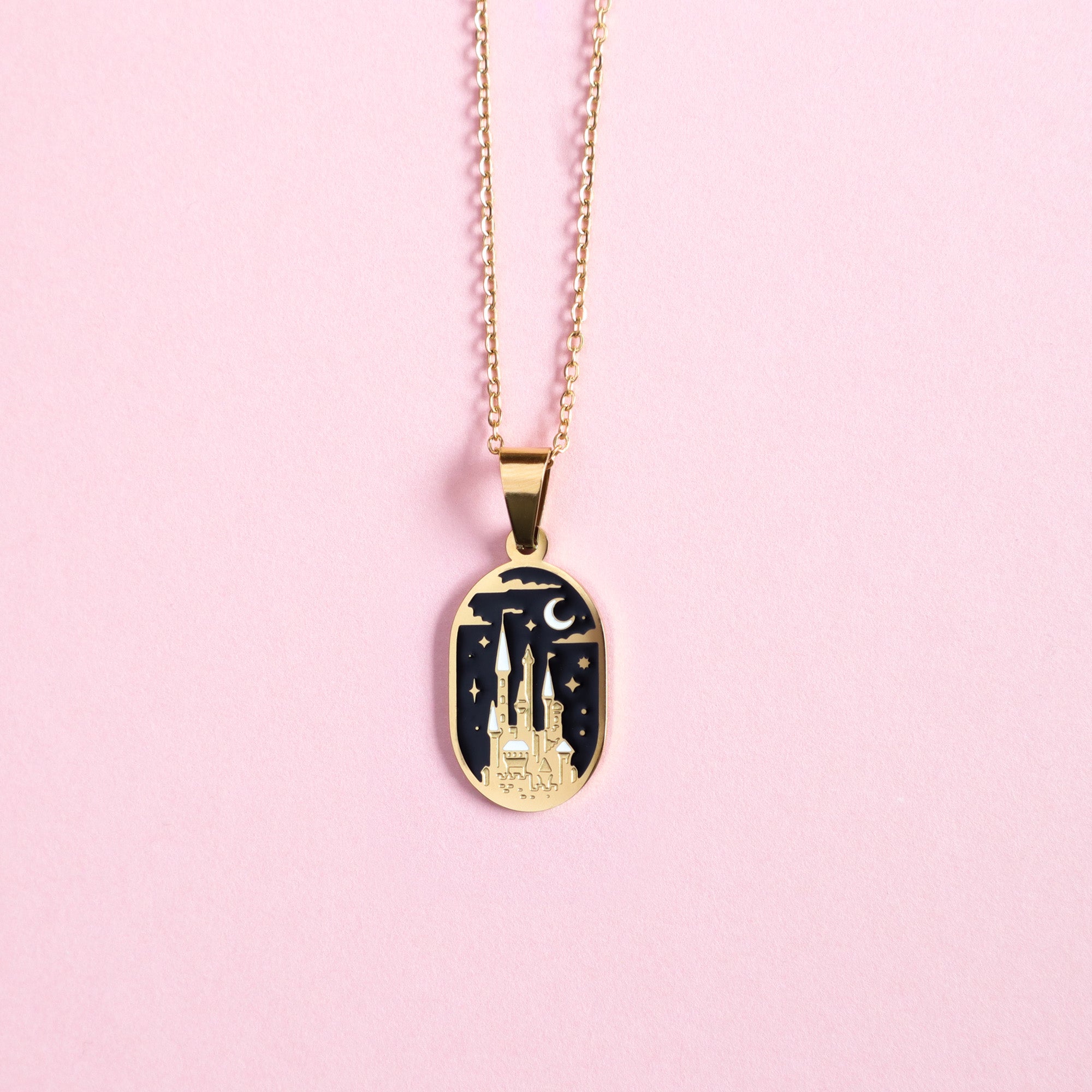 The Papio Press Moonlit Castle Enamel Necklace features a gold oval pendant with a castle, stars, and crescent moon on a dark background, beautifully displayed on a light pink surface.