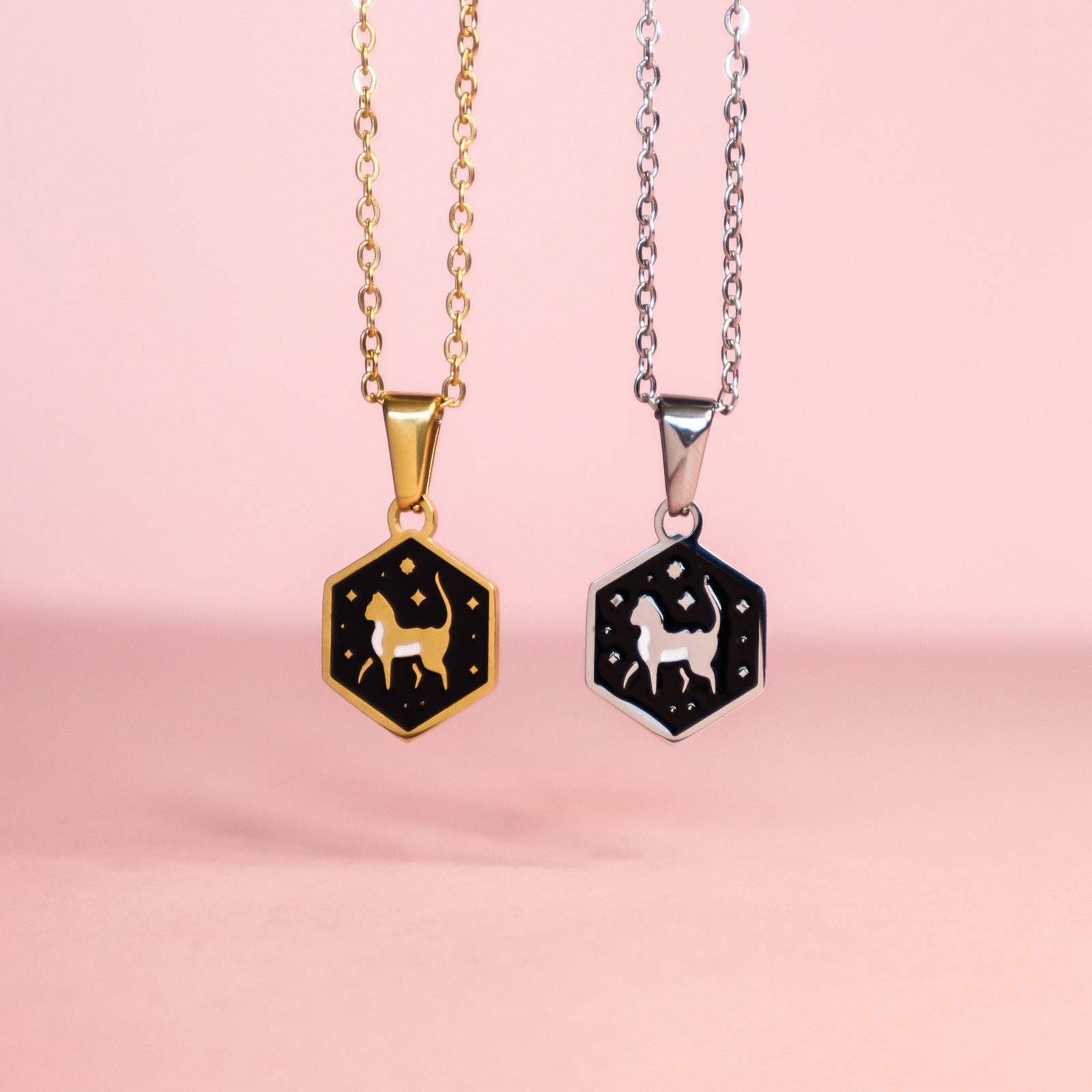 Two Mini Hexagon Cat Pendants by Papio Press—one gold, one silver—on stainless steel chains with white cat silhouettes and stars on black enamel, displayed on a soft pink background.