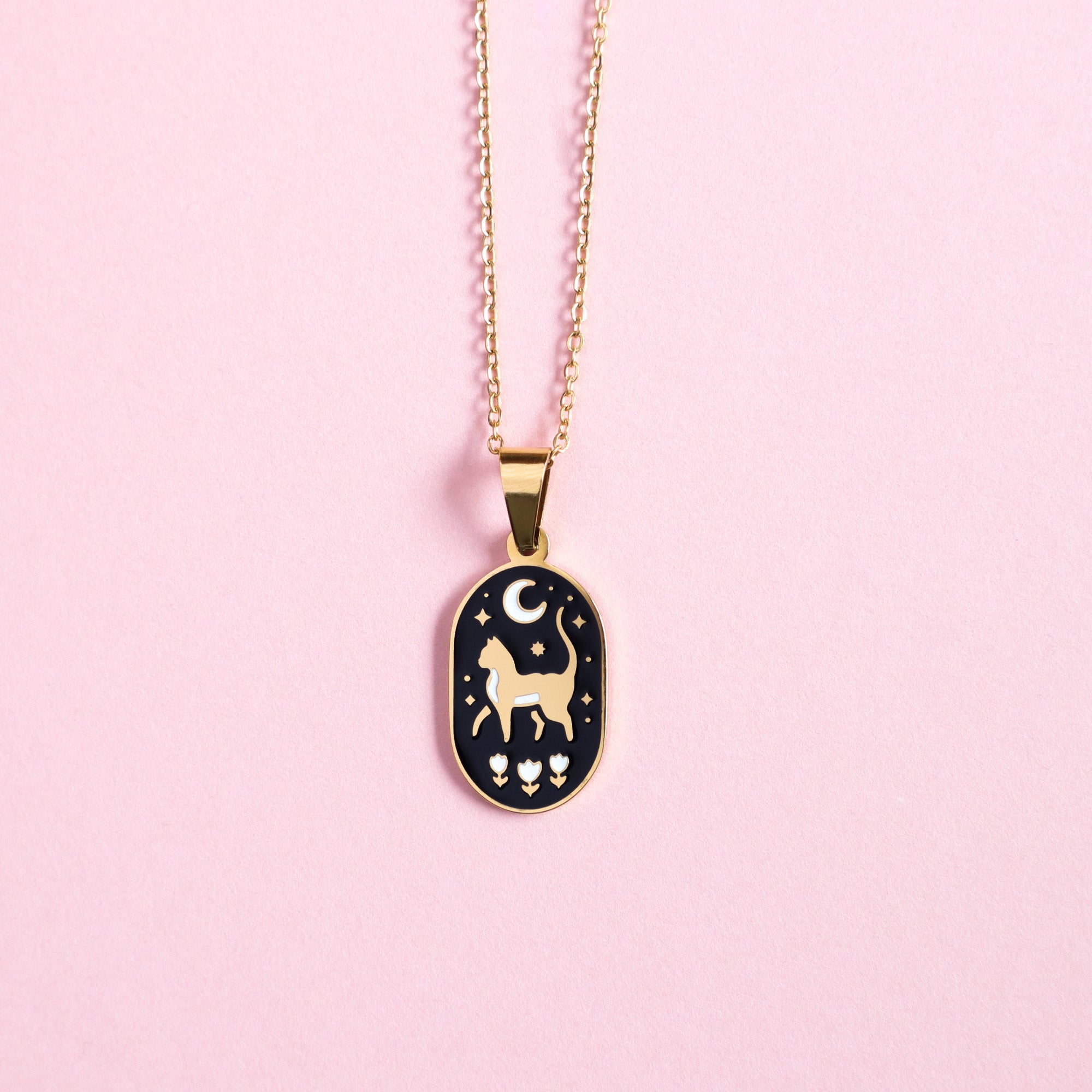 The Papio Press Cat & Moon Enamel Necklace features an oval pendant with a white cat, flowers, stars, and a crescent moon on black enamel, all set on a durable stainless steel base and displayed on a light pink surface.