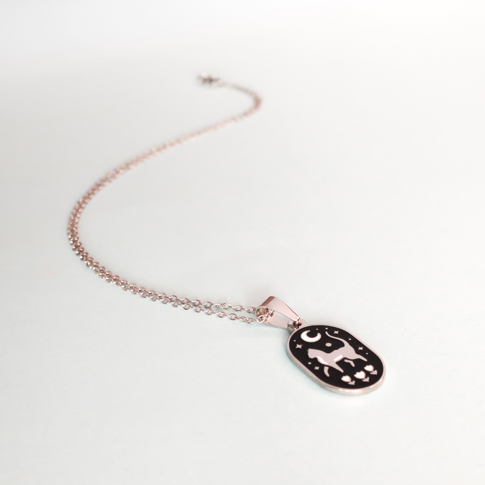 The Cat & Moon Enamel Necklace by Papio Press features a delicate gold chain and a black oval pendant with a white cat, stars, and a crescent moon, beautifully displayed on a light background.