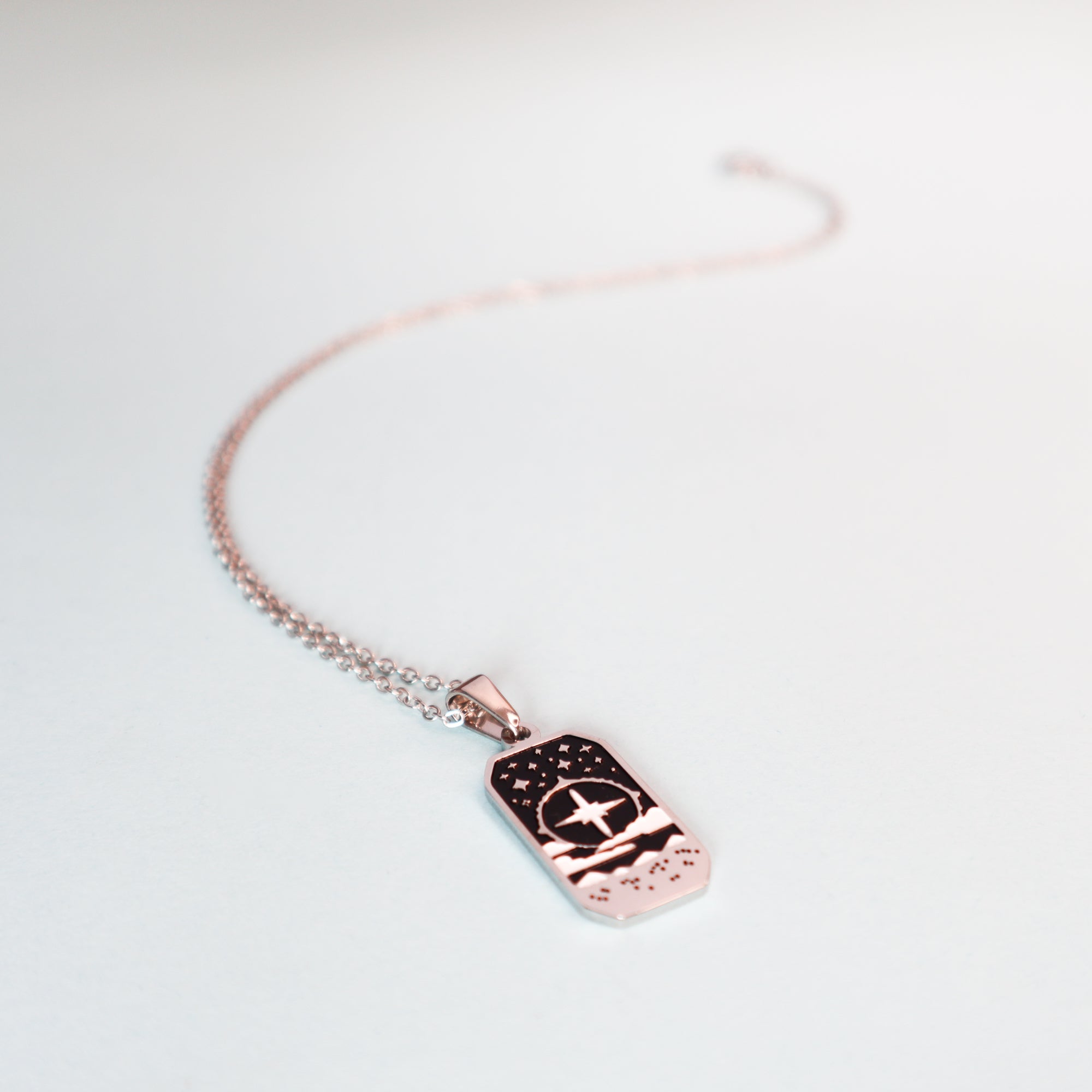 The Cleric Pendant Enamel Necklace by Papio Press features a rose gold rectangular pendant engraved with stars and mountains, adding a touch of fantasy jewelry to any collection. Displayed on a white background.
