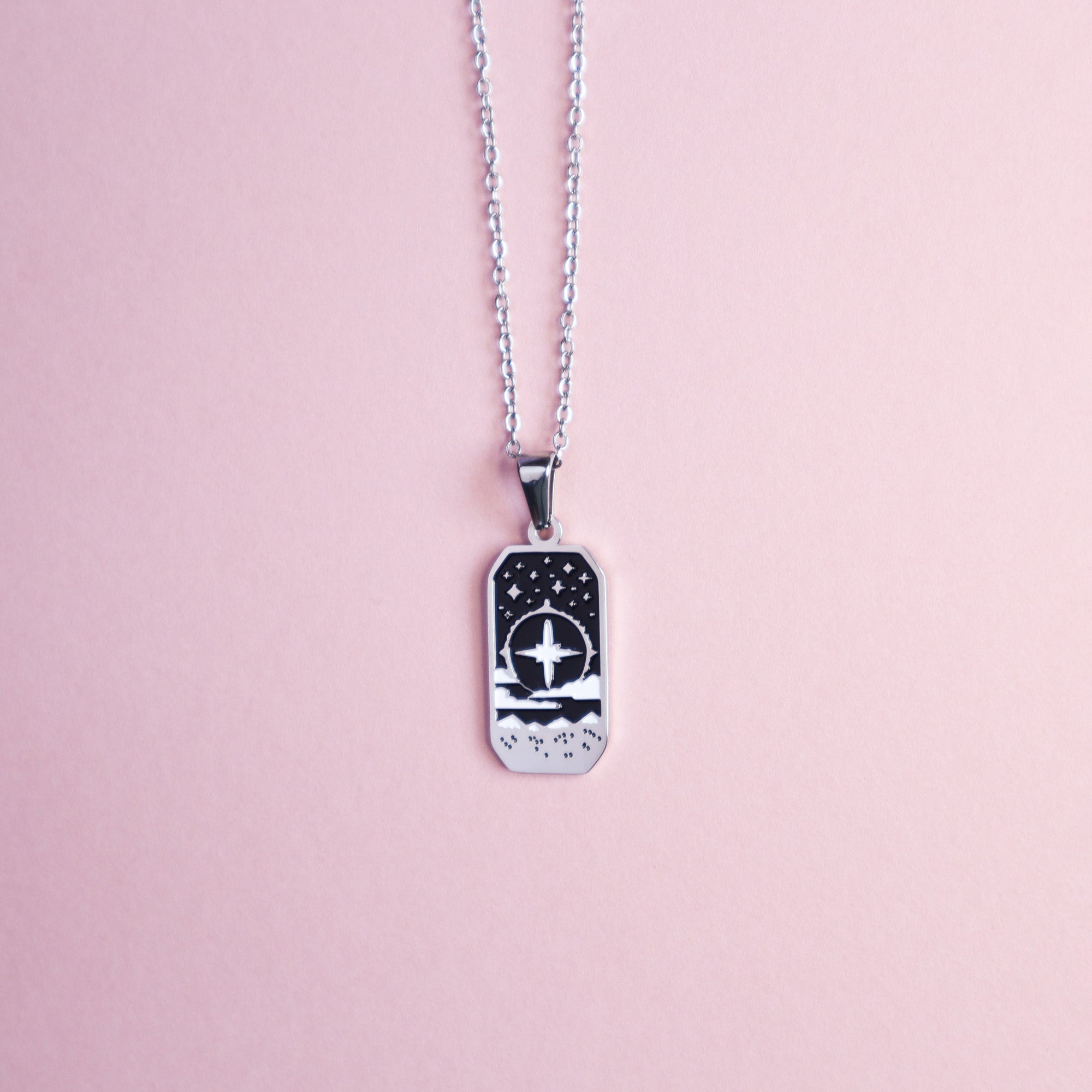 The Cleric Pendant Enamel Necklace by Papio Press features a silver recycled chain and a rectangular pendant with a black and white cross, waves, and stars—fantasy jewelry ideal for tabletop gamers. Shown on a light pink background.