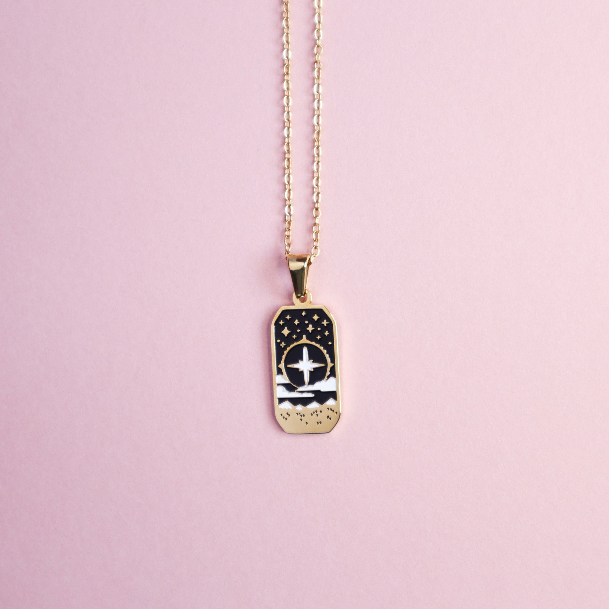 The Cleric Pendant Enamel Necklace by Papio Press features a gold rectangular charm with an engraved cross, stars, and waves—ideal fantasy jewelry for tabletop game fans—shown on a pale pink background.