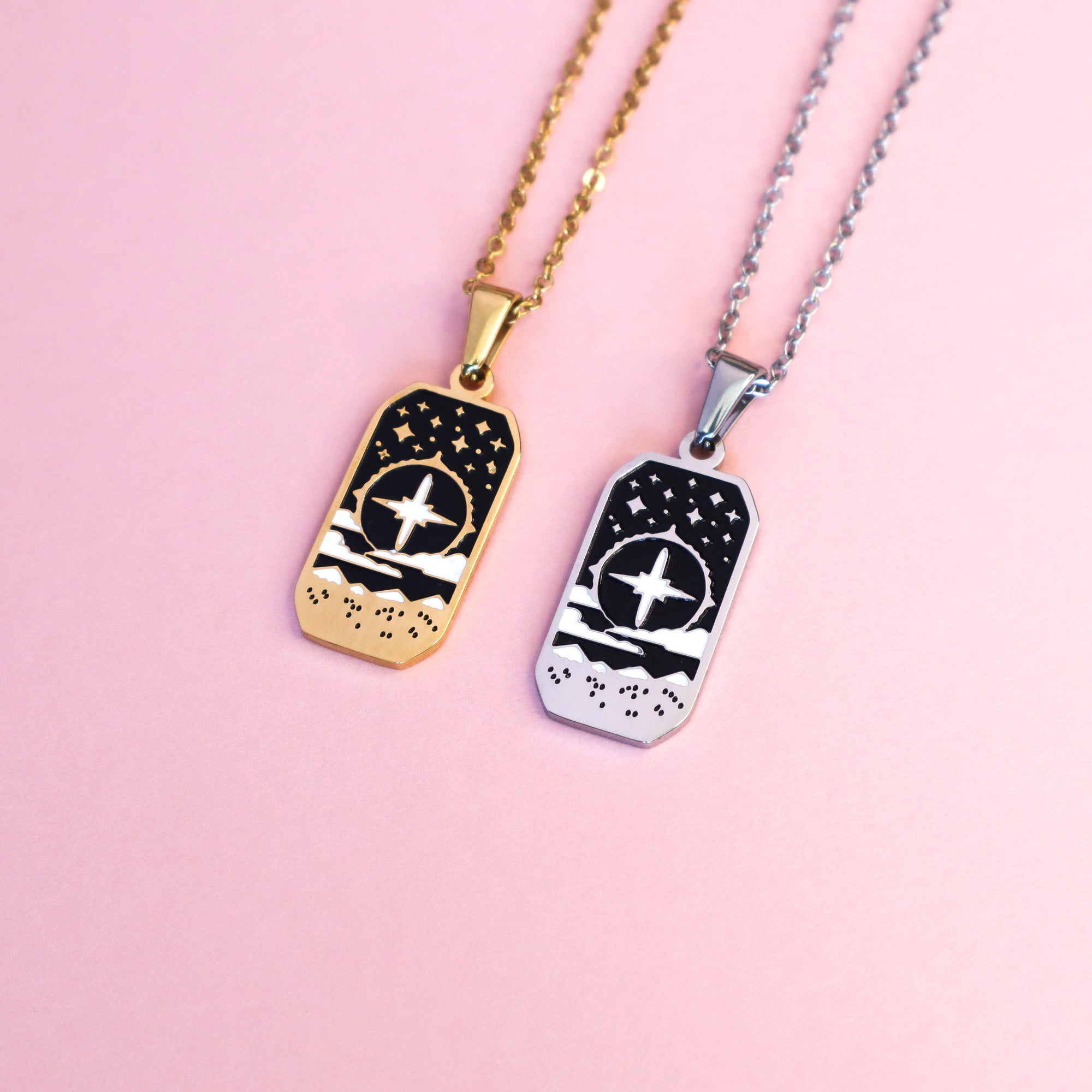 Two rectangular Cleric Pendant Enamel Necklaces by Papio Press—one silver, one gold—each featuring mountains, stars and clouds displayed on a light pink surface. Ideal for tabletop game lovers.