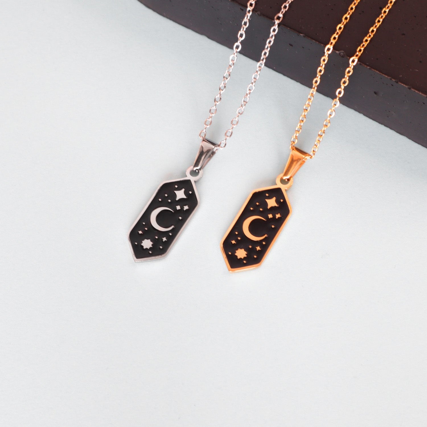 Two Papio Press Crescent Moon Pendant Enamel Necklaces with hexagonal black pendants—featuring a crescent moon and star—are shown on silver and gold plated chains, displayed on a light surface next to a dark object.