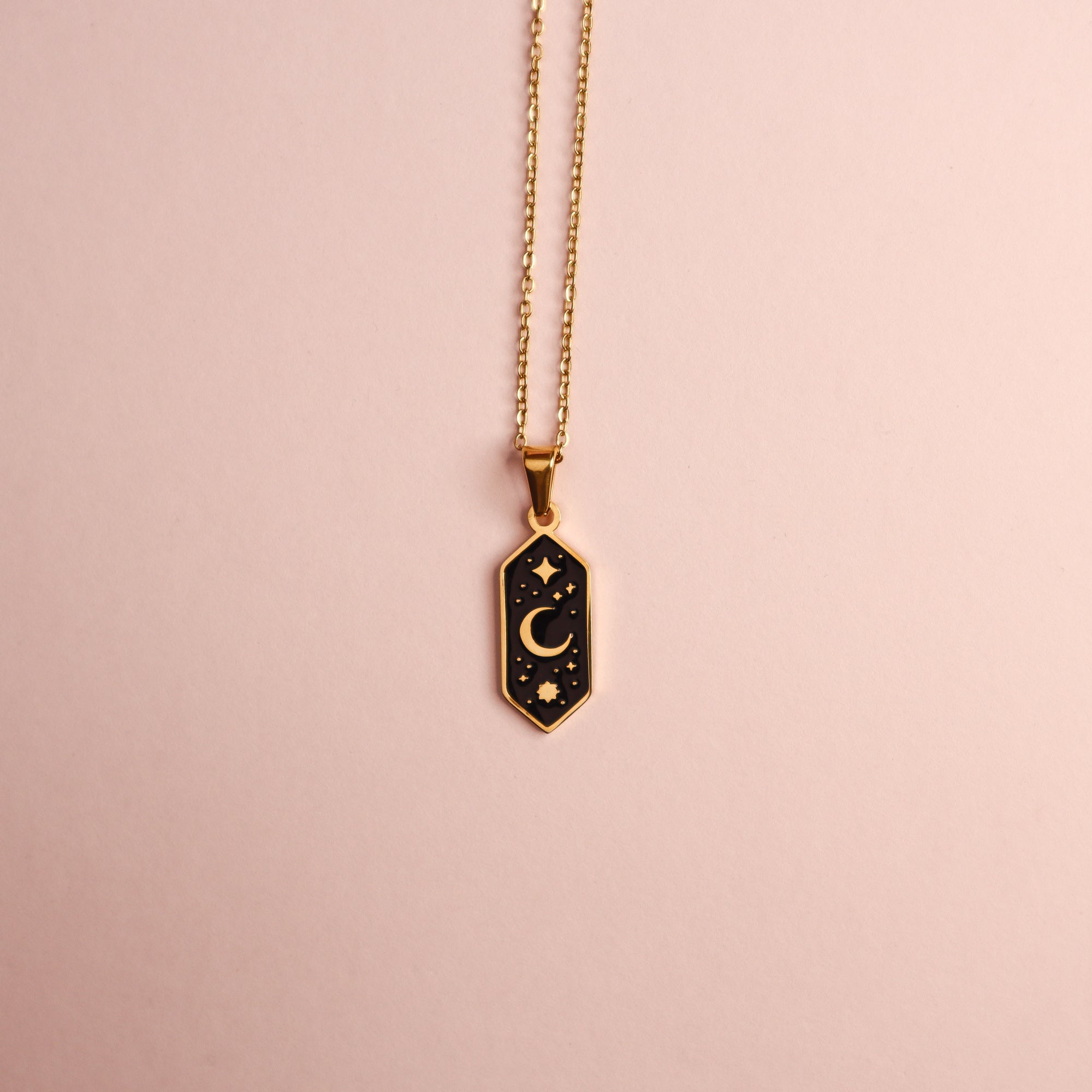 The Crescent Moon Pendant Enamel Necklace by Papio Press features a gold plated chain and a rectangular black enamel charm with a crescent moon and stars, displayed on a light pink background.