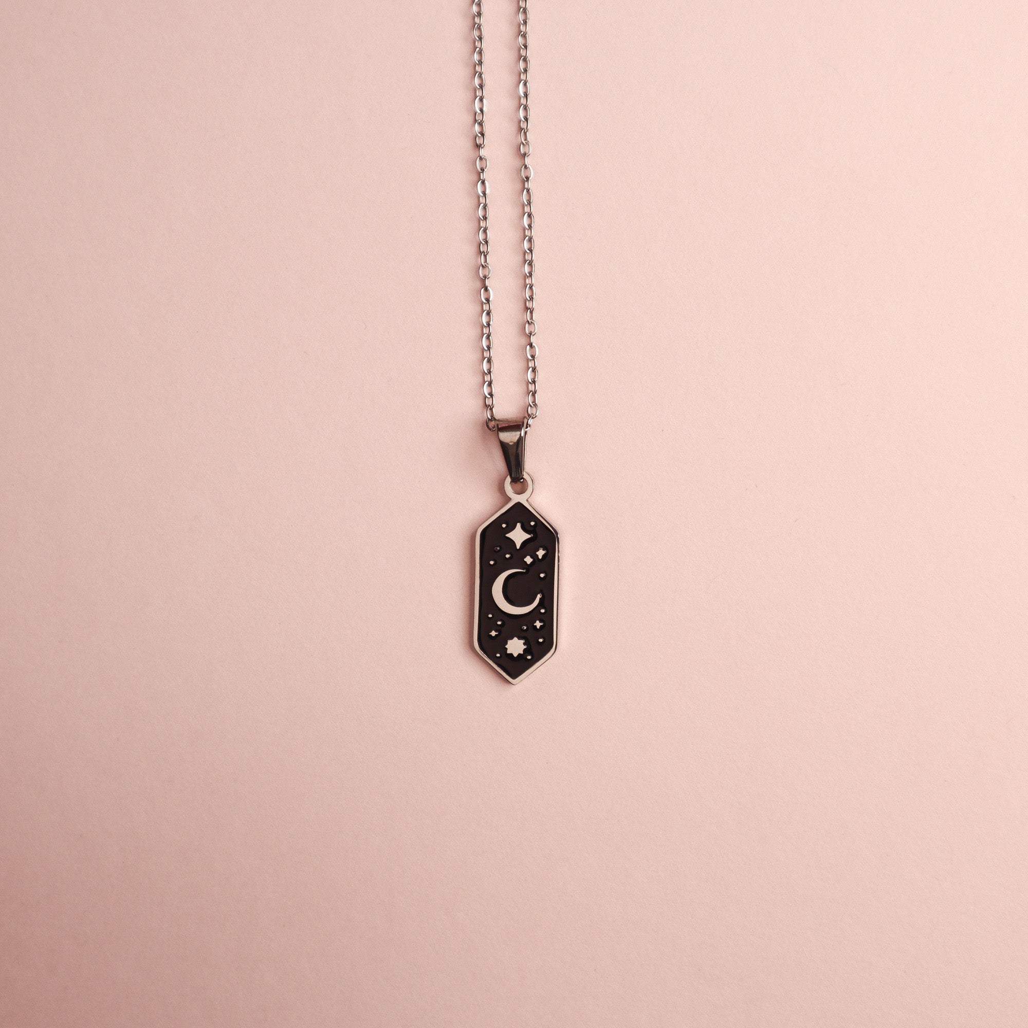 The Crescent Moon Pendant Enamel Necklace by Papio Press features a hexagonal black pendant with a gold crescent moon and star, displayed on a light pink background.