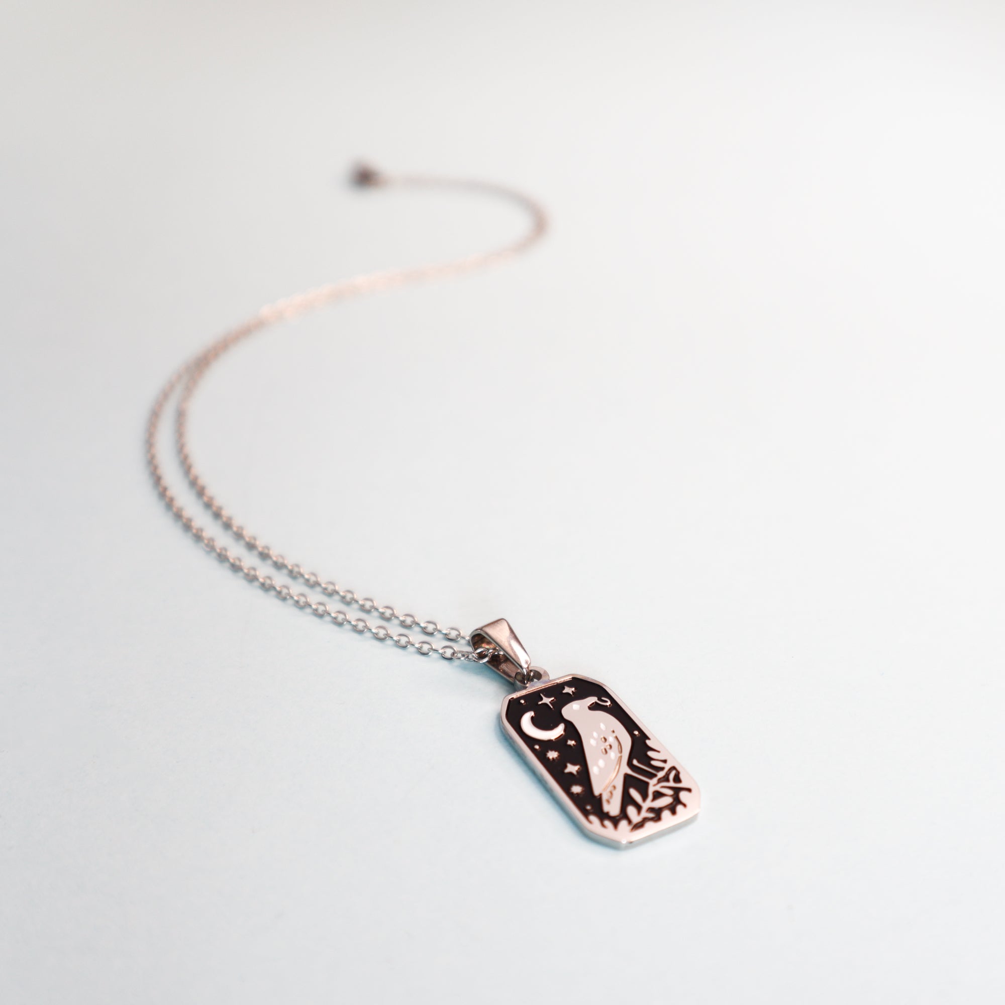 The Crow and the Ring Pendant Enamel Necklace by Papio Press features a rose gold rectangular pendant with an engraved bird, crescent moon, and stars. The curved chain rests on a white surface—ideal for fans of fantasy jewelry.
