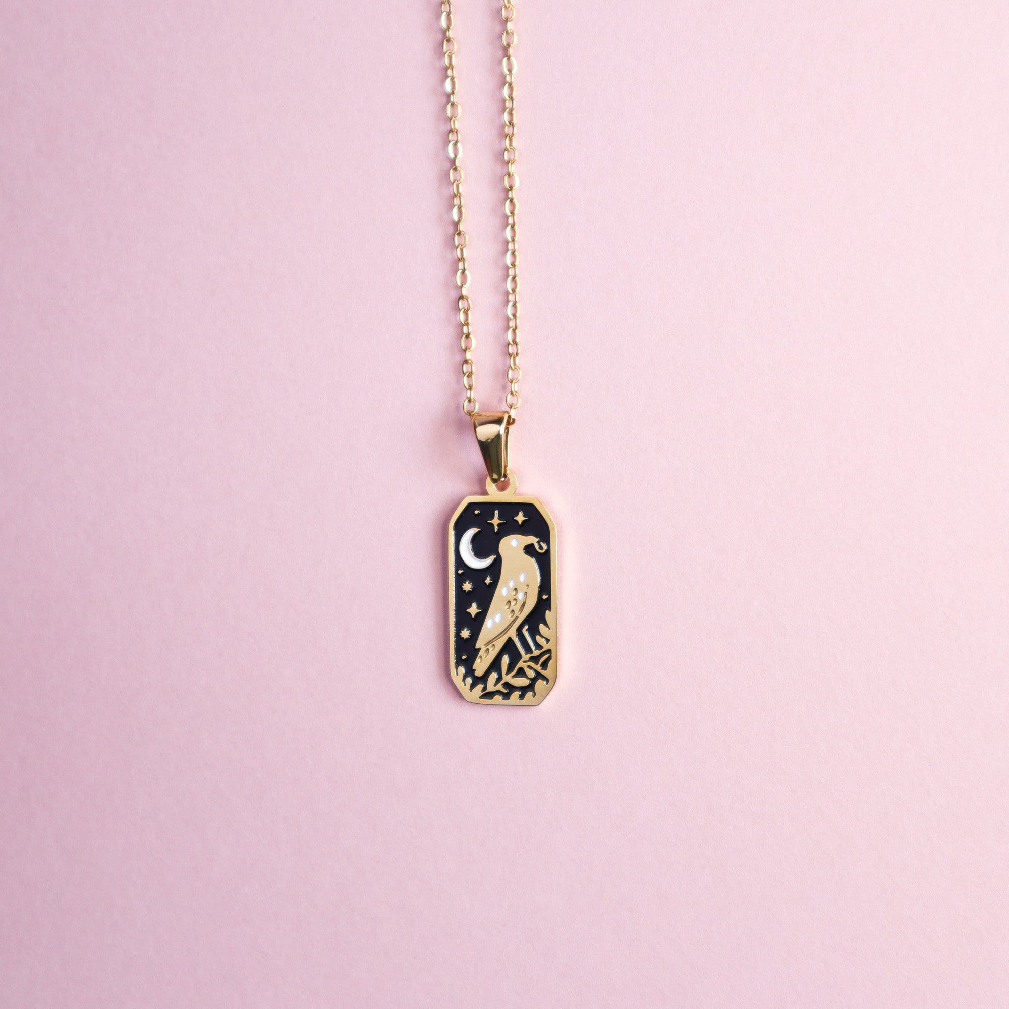 The Crow and the Ring Pendant Enamel Necklace by Papio Press features a gold rectangular charm with an engraved bird, crescent moon, and stars on a black background, beautifully displayed on a light pink surface.