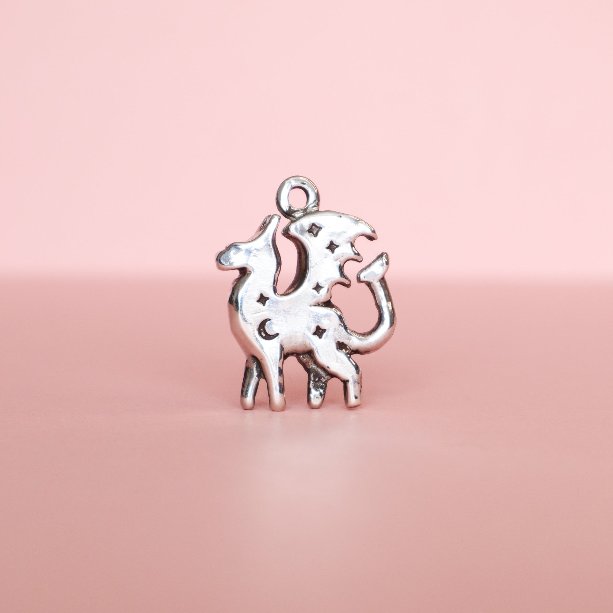 The Silver Lunar Dragon Pendant by Papio Press is a unique handmade jewelry piece, crafted from recycled silver and featuring star and moon cutouts, shown against a plain pink background.