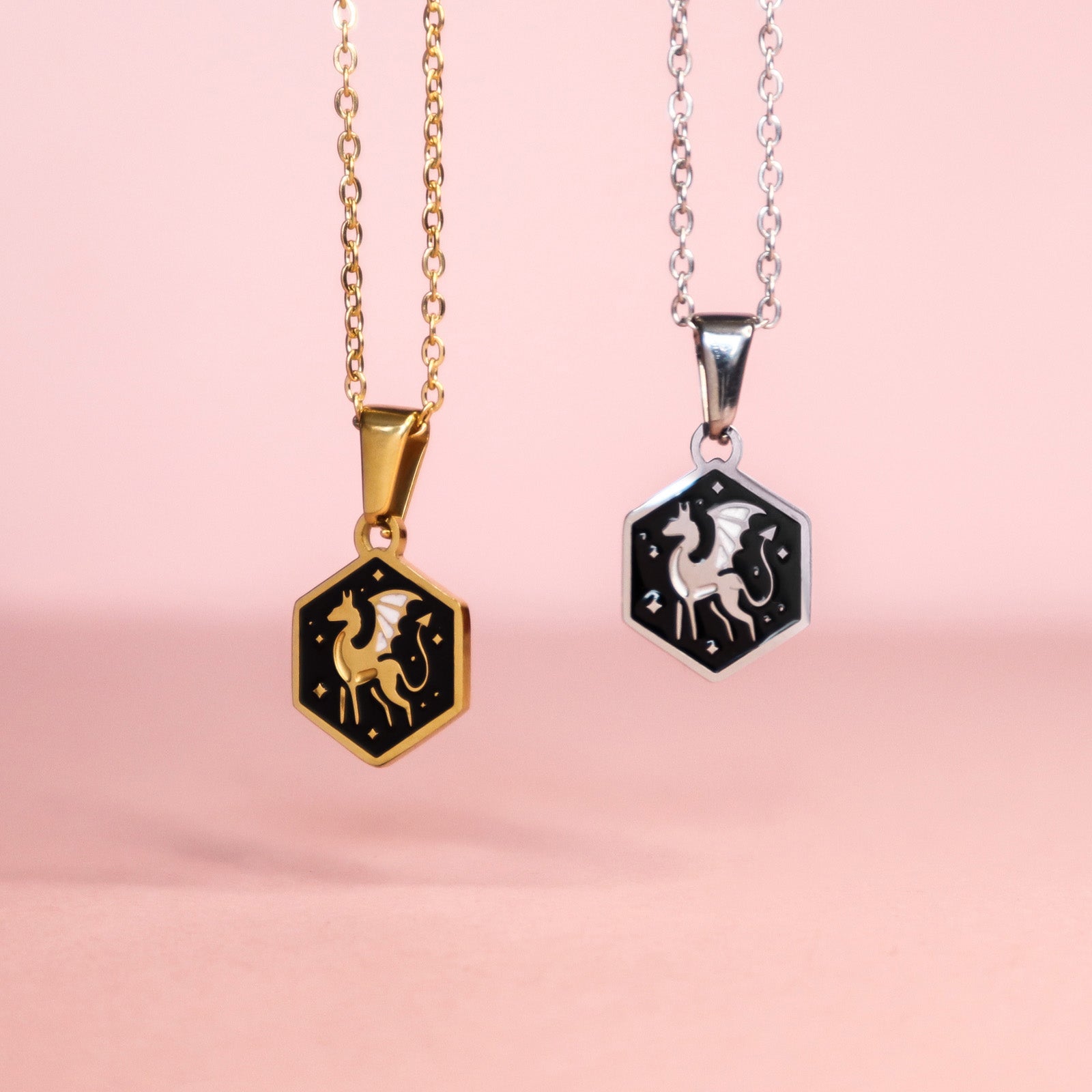Two mini hexagon pendants on chains by Papio Press—one gold, one silver—each feature a dragon design on black enamel, creating a whimsical necklace set called the Mini Hexagon Dragon Pendant. Displayed against a soft pink background.