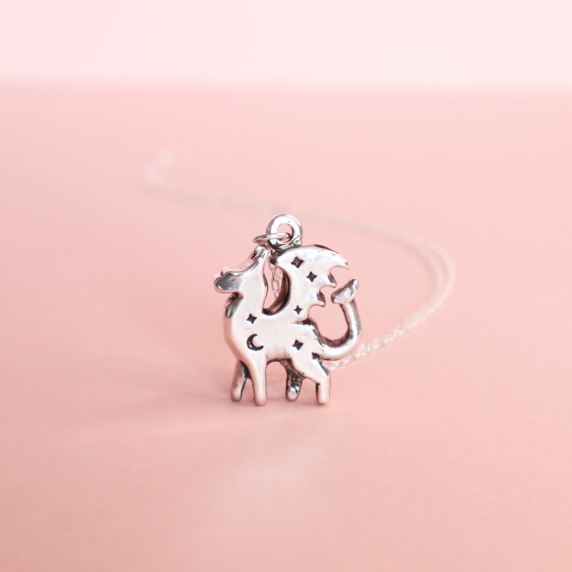 The Silver Lunar Dragon Pendant by Papio Press gleams against a soft pink background—a whimsical handmade silver piece featuring a dragon design, perfect for those who love dreamy jewelry styles.