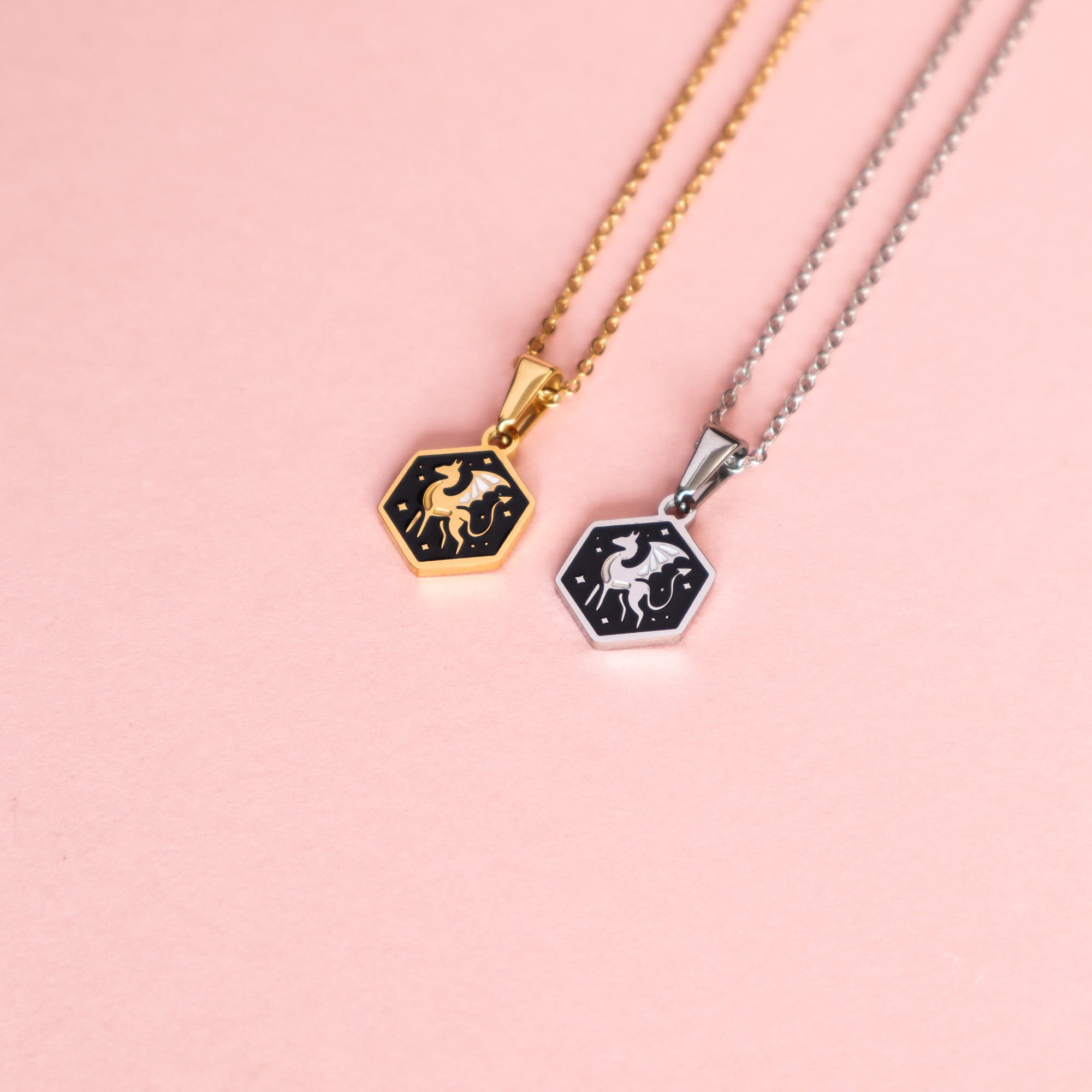 Two Mini Hexagon Dragon Pendants by Papio Press rest on a pink background—one with a gold chain, one with a silver stainless steel chain. Each features a white dragon design on black, adding fantasy-inspired charm to your look.