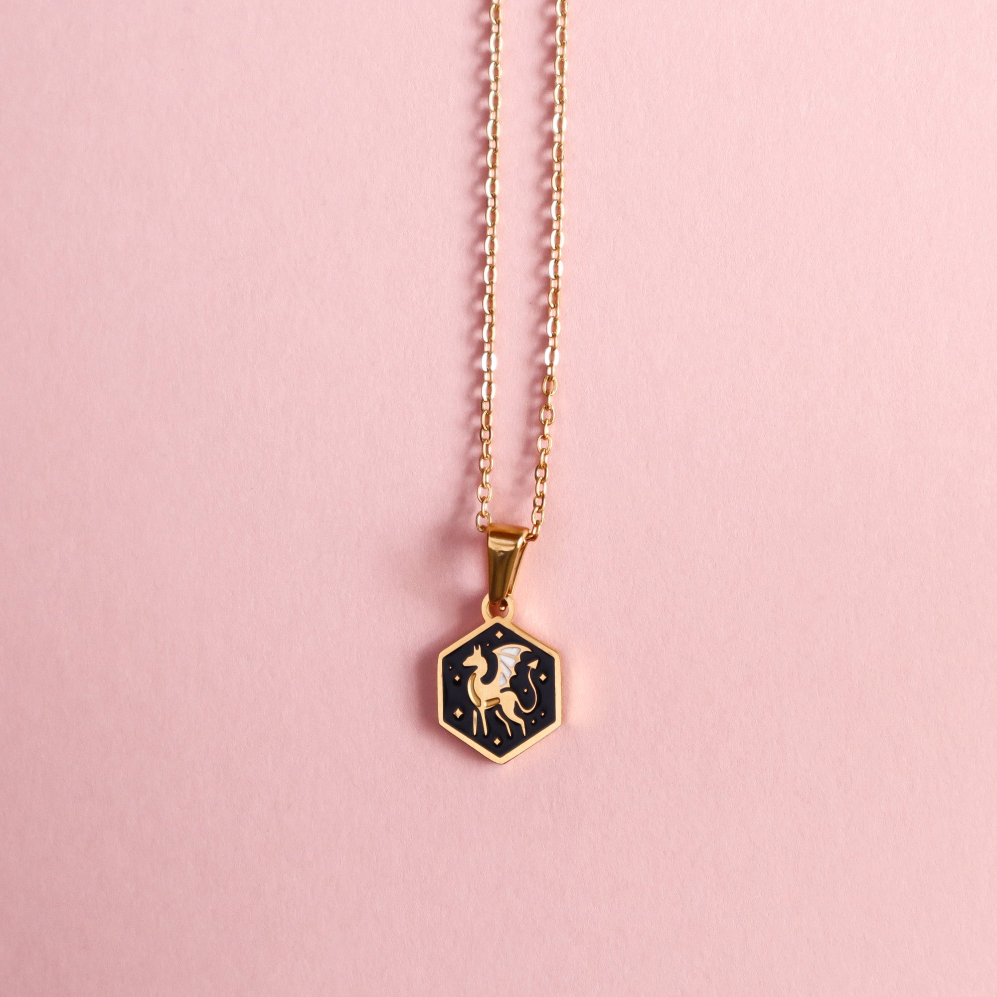 The Papio Press Mini Hexagon Dragon Pendant is a gold enamel necklace with a dragon design, displayed on a light pink surface.