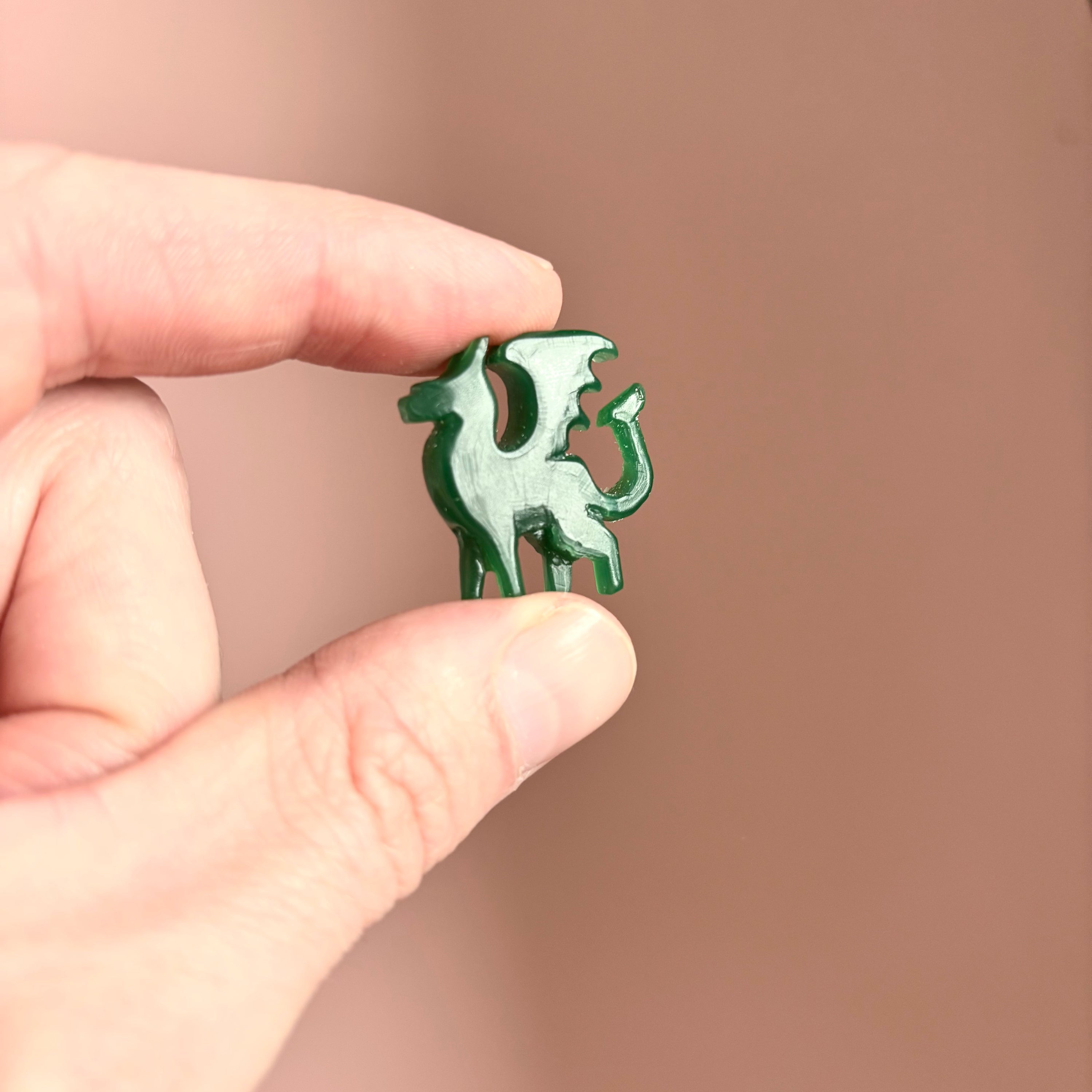 A hand holds a small, green, dragon-shaped wax carving of the Silver Lunar Dragon Pendant by Papio Press, set against a plain, light brown background.