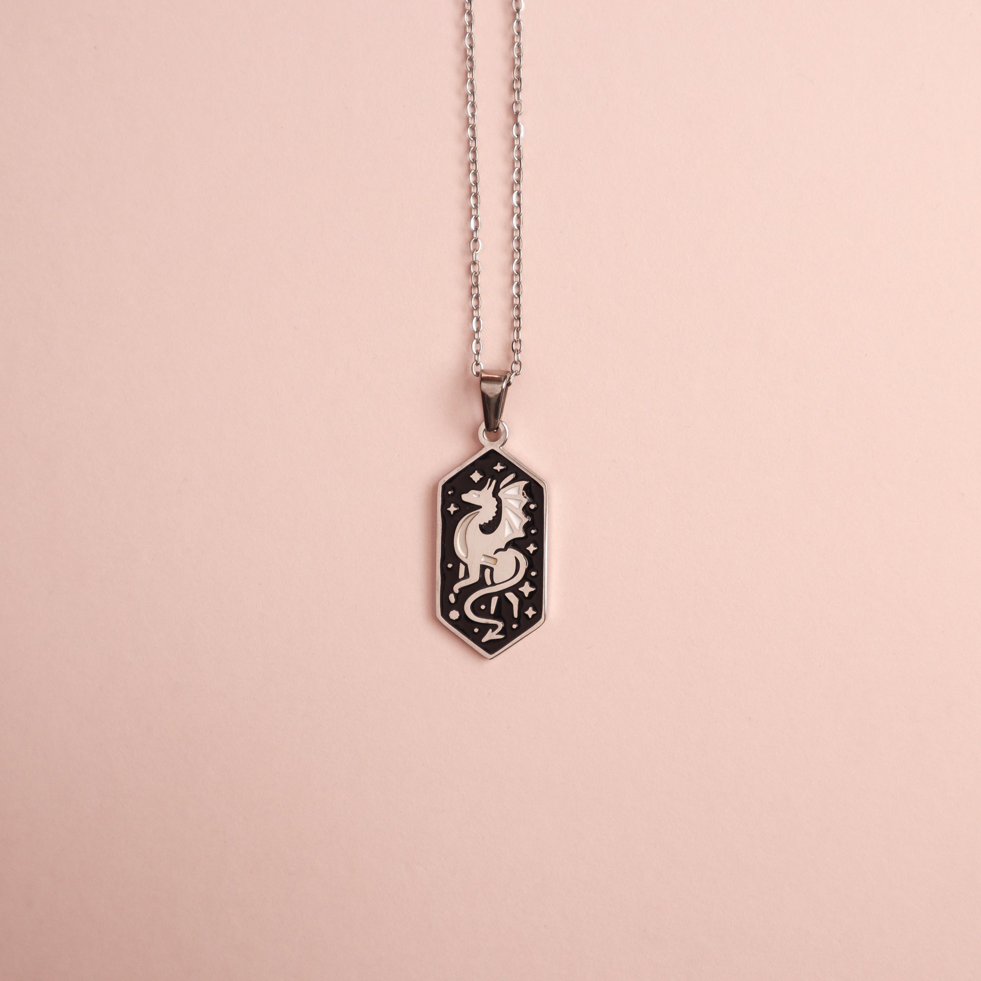 The Papio Press Dragon Pendant Enamel Necklace features a hexagonal charm with an engraved dragon and stars, set on a pale pink background. Pair it with an enamel piece or 18K gold plated accents for extra elegance.