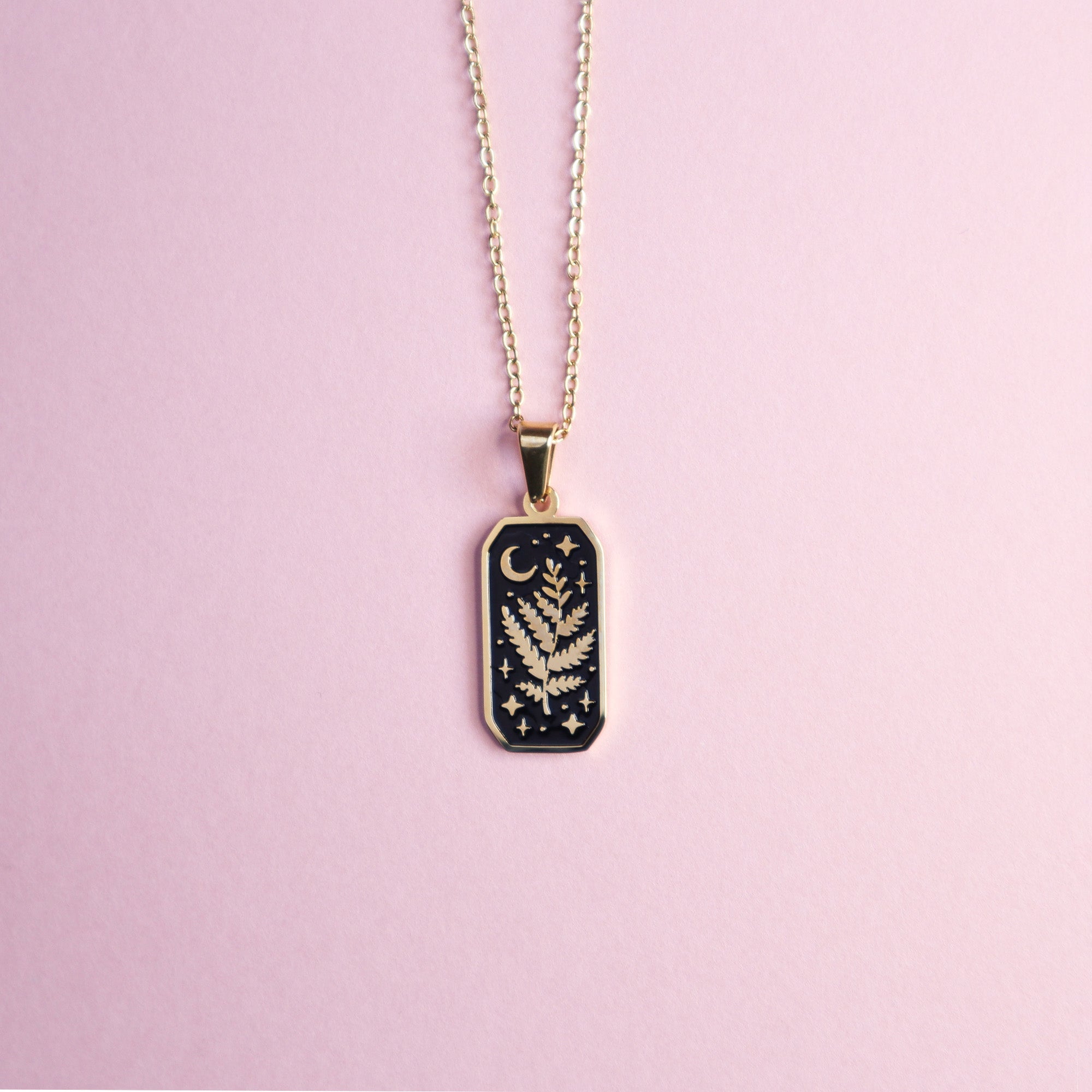 The Druid Pendant Enamel Necklace by Papio Press features a rectangular black enamel pendant with a gold botanical motif, crescent moon, and stars on a light pink background for a whimsical touch.