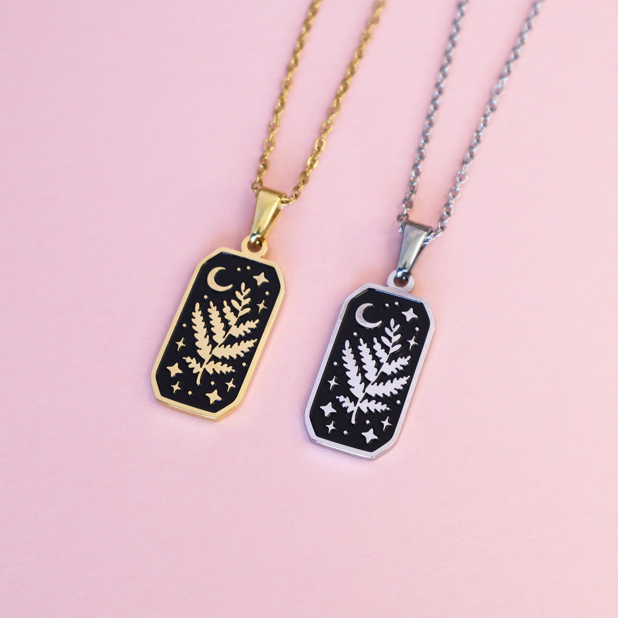 The Druid Pendant Enamel Necklaces by Papio Press feature gold and silver stainless steel chains with a white botanical design, crescent moon, and stars on black—an enchanting fantasy accessory shown on a pastel pink surface.