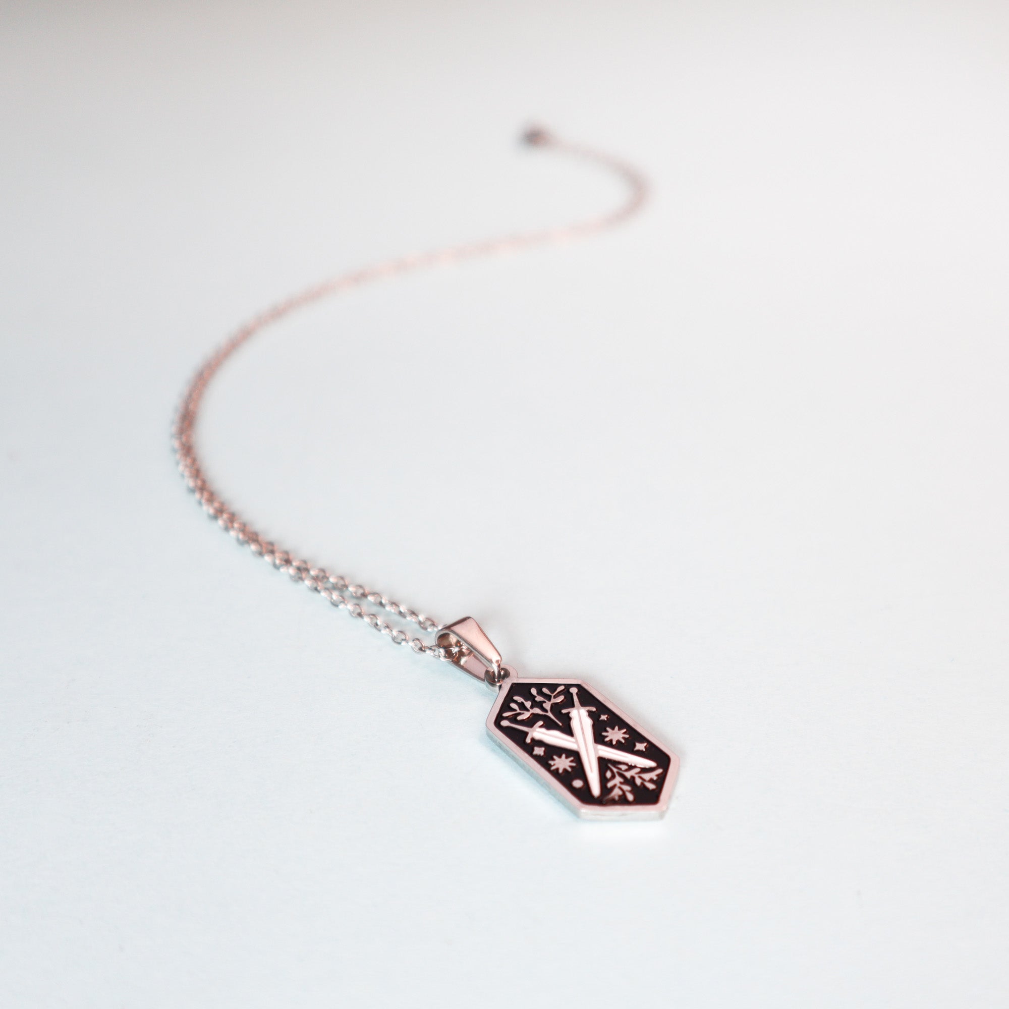 The Papio Press Dual Swords Pendant Enamel Necklace features a rose gold hexagonal pendant with engraved swords and star details, perfect for fantasy lovers, shown on a white background.