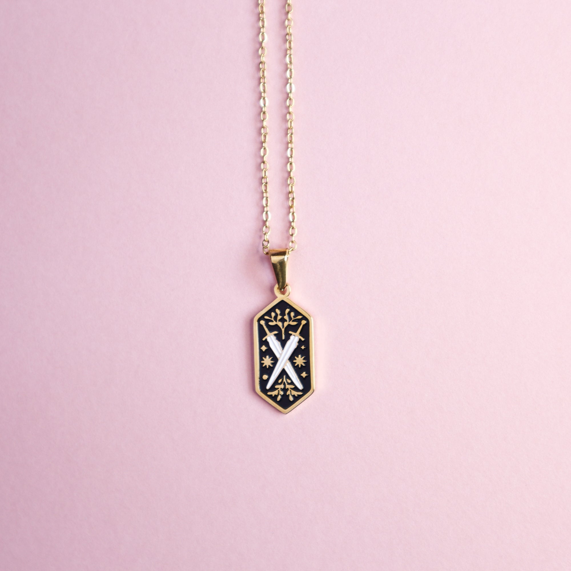 Fantasy fans will love the Papio Press Dual Swords Pendant Enamel Necklace, featuring a gold hexagonal pendant with black enamel, a white X, and whimsical floral accents—perfect for tabletop gamers. Shown on a pink background.