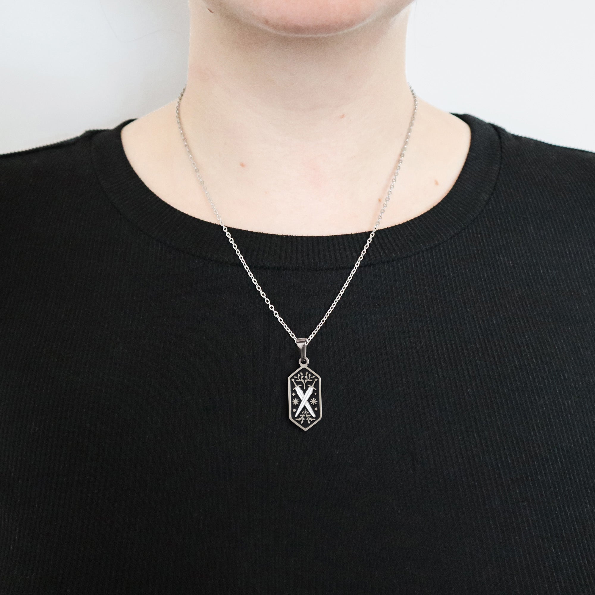 A person wears a plain black ribbed shirt and the Papio Press Dual Swords Pendant Enamel Necklace, featuring a black pendant with a white geometric design. The photo is cropped to show only the neck and upper chest. Perfect for fantasy lovers.