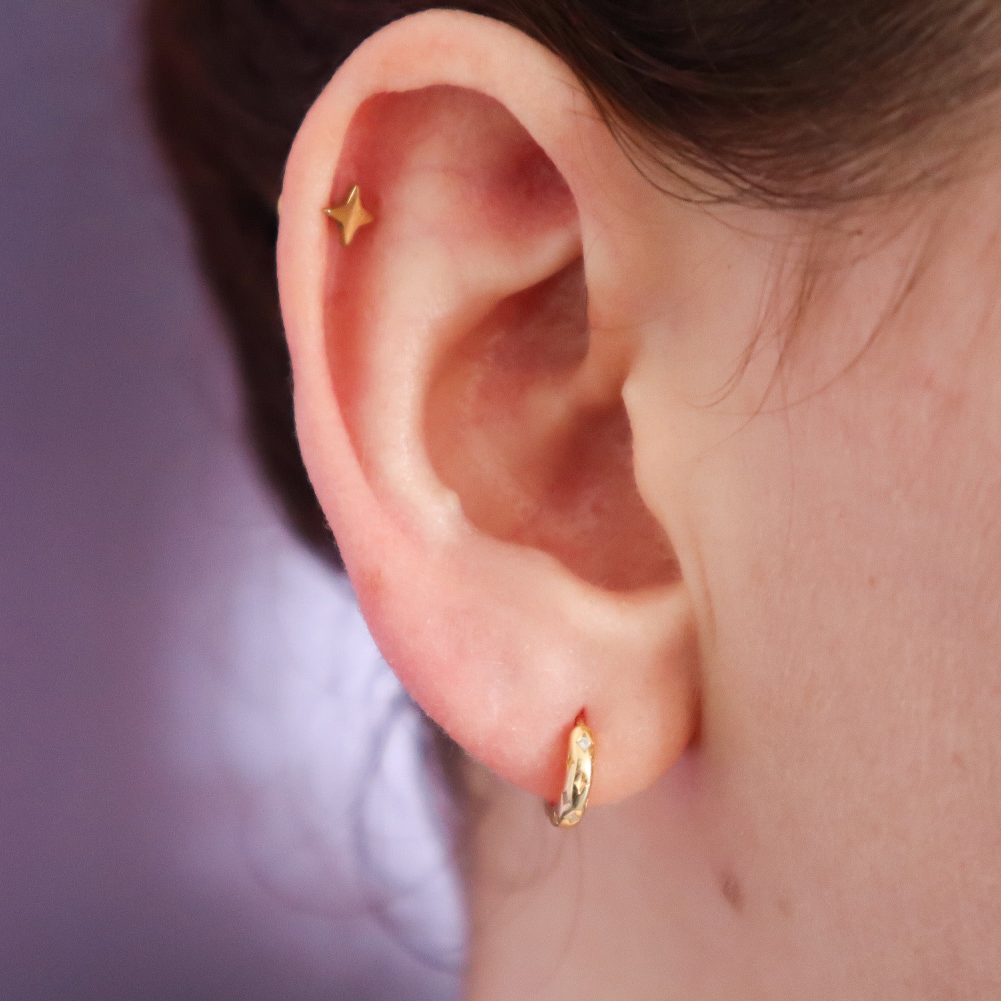 A close-up shows two Papio Press Star Huggie Hoop Earrings: a small gold star stud in the upper cartilage and a delicate gold huggie hoop in the earlobe, with a softly blurred background.
