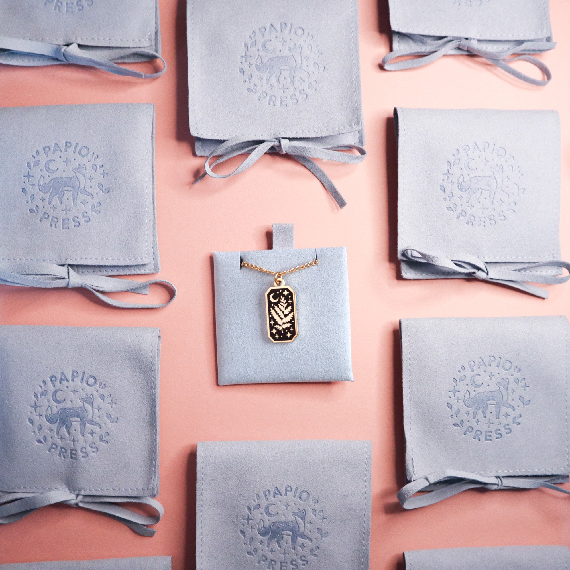 The Bard Pendant Enamel Necklace by Papio Press, featuring a celestial design, is shown on a light blue pouch amid other branded pouches with an animal logo—an enchanting accessory for fantasy lovers, displayed on a pink surface.