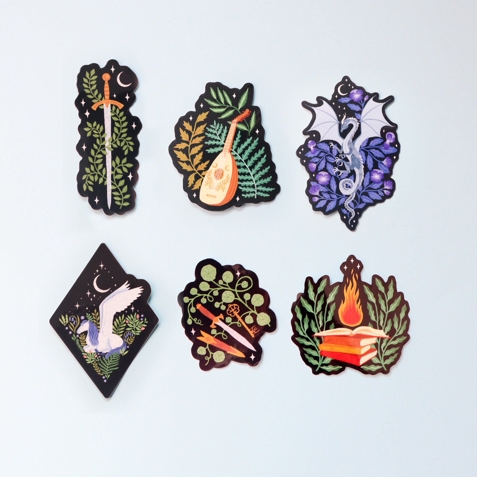 The Fantasy Sticker Collection by Papio Press includes six glossy, UV-protected vinyl stickers featuring a sword with ferns and moon, a lute with leaves, a silver dragon, white bird, crossed arrows with clover, and flaming book.