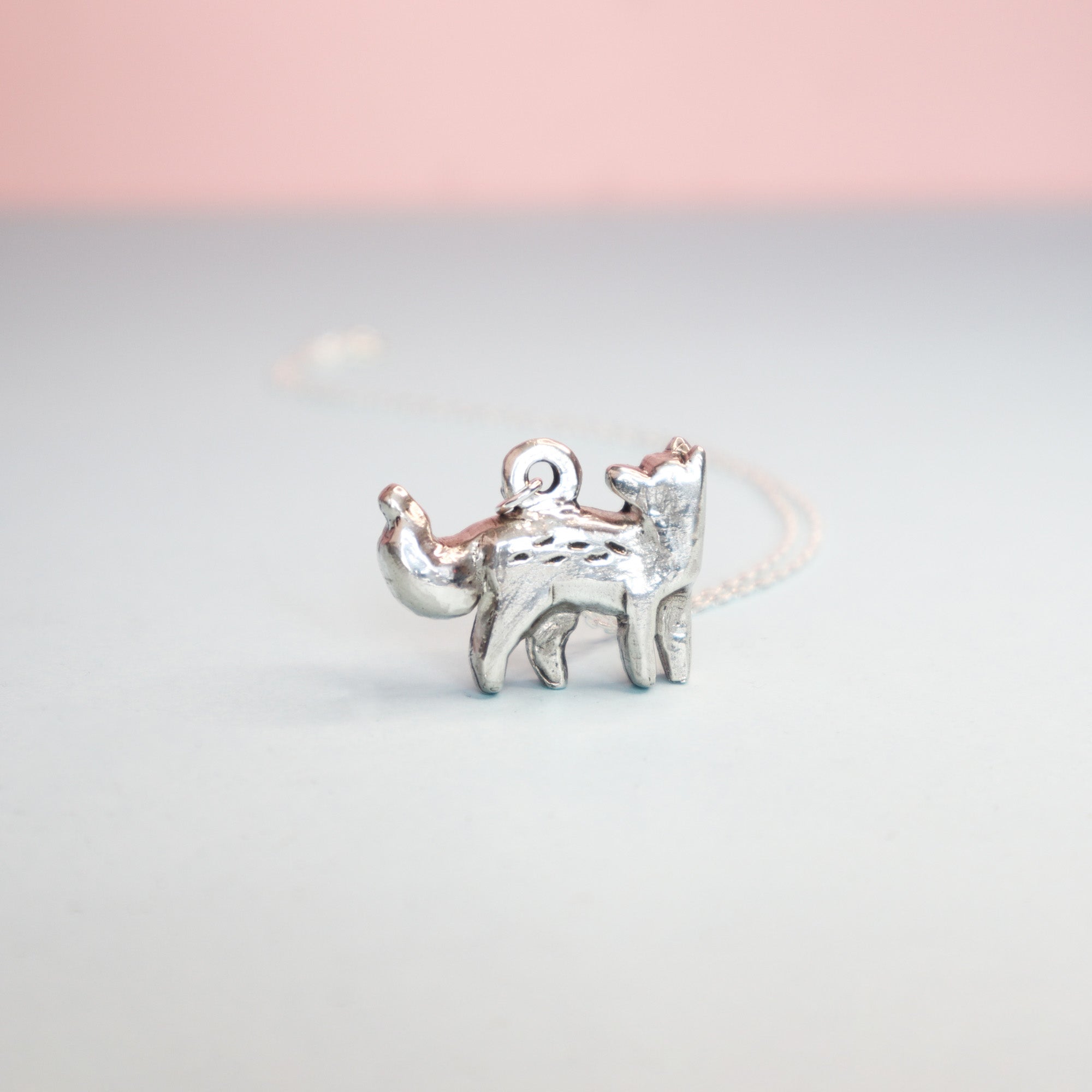 The Papio Press Silver Fox Pendant, a handmade silver jewelry piece with enamel detail, is shown on a light surface against a softly blurred pink and blue background.