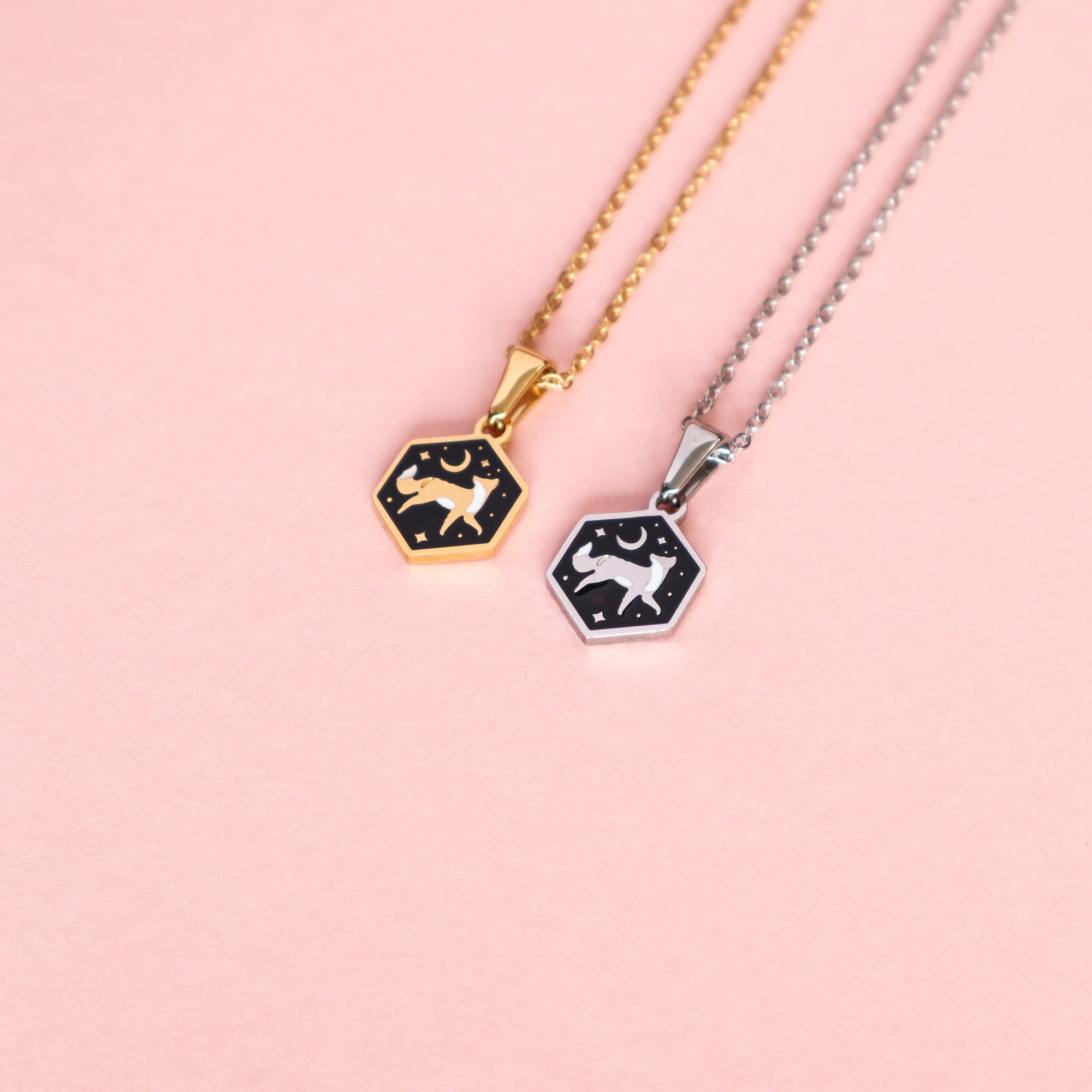 Two hexagon-shaped enamel necklaces from Papio Press—Mini Hexagon Fox Pendants on gold and silver stainless steel chains—feature a fox, stars, and a crescent moon on black backgrounds, displayed on a light pink surface.