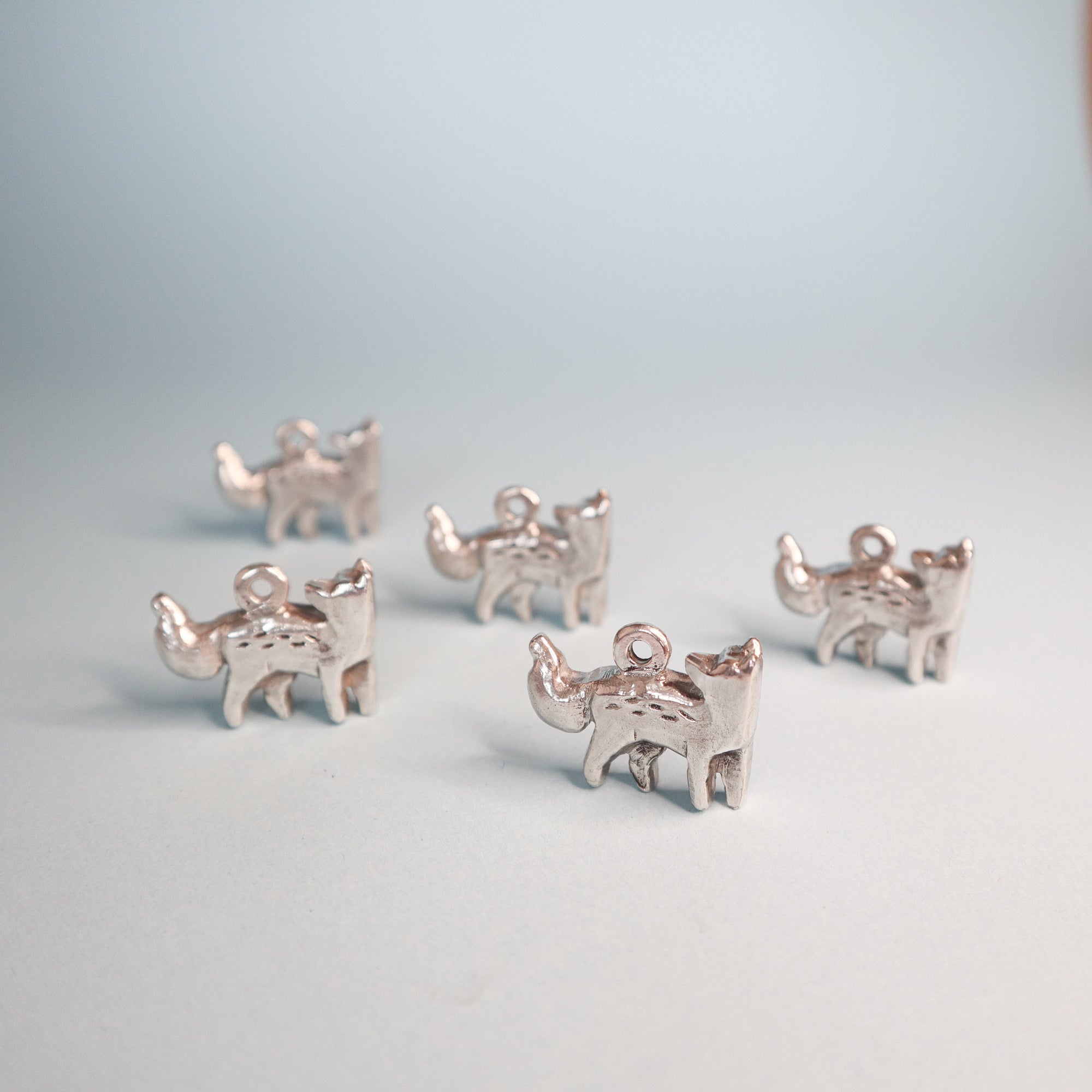 Five small Silver Fox Pendants by Papio Press are arranged on a pale surface with a soft blue background. Each features a loop at the top, ideal for pairing with a sterling silver chain or adding to your favorite accessories.