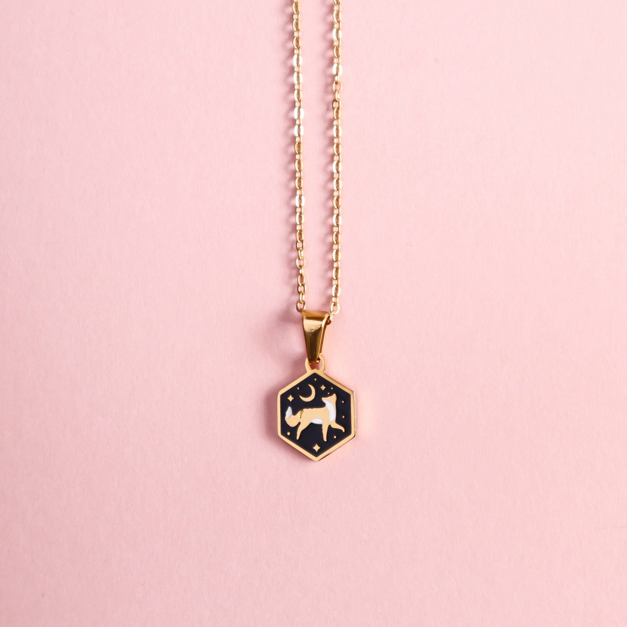The Papio Press Mini Hexagon Fox Pendant is a gold stainless steel chain necklace with a hexagonal black enamel fox charm featuring a white animal and star design, shown on a light pink background.