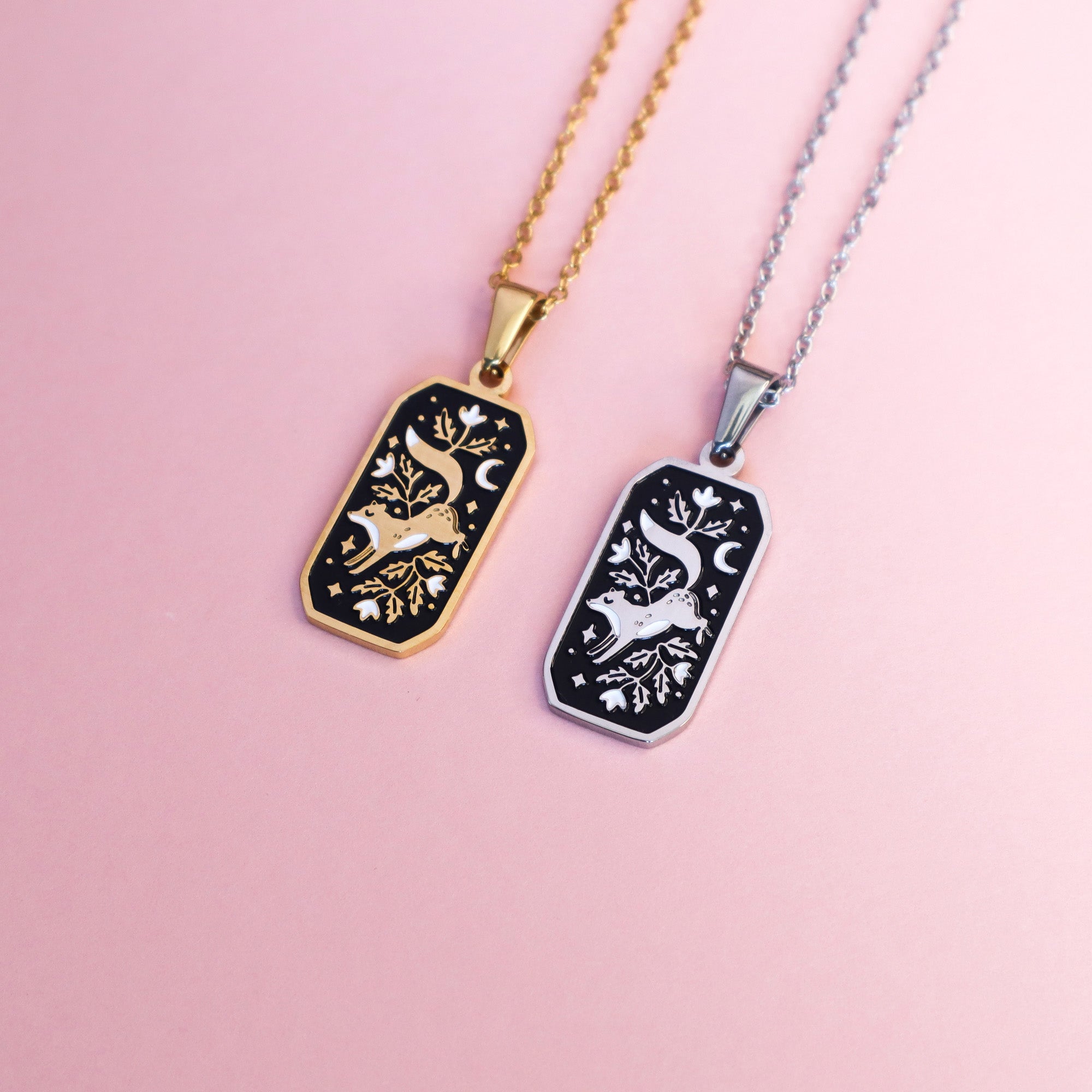 Two Papio Press Floral Fox stainless steel necklaces, one in gold and one in silver. The pendants are rectangular and feature a leaping fox surrounded by florals, stars and a moon. Each feature black and white enamel details taken a light pink surface.