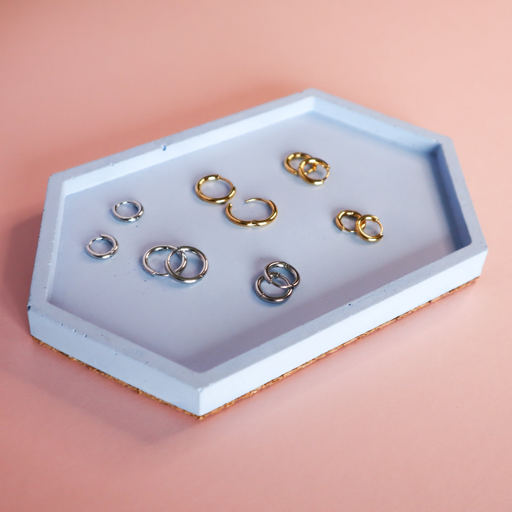 A selection of gold and silver huggie hoop earrings on a blue tray. The tray is on a pink background.