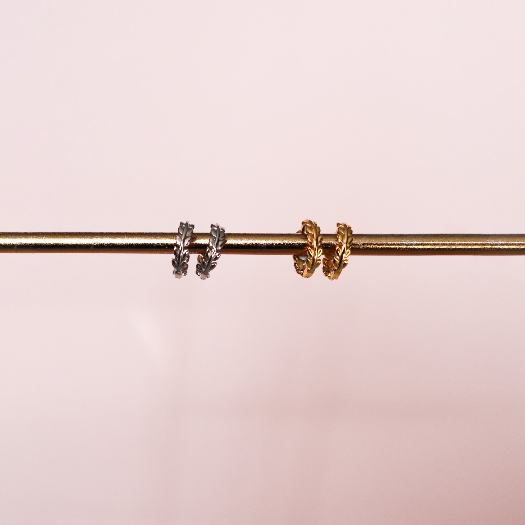 Two pairs of leafy laurel hoops, one gold and one silver attached to a golden pole on a pink background.
