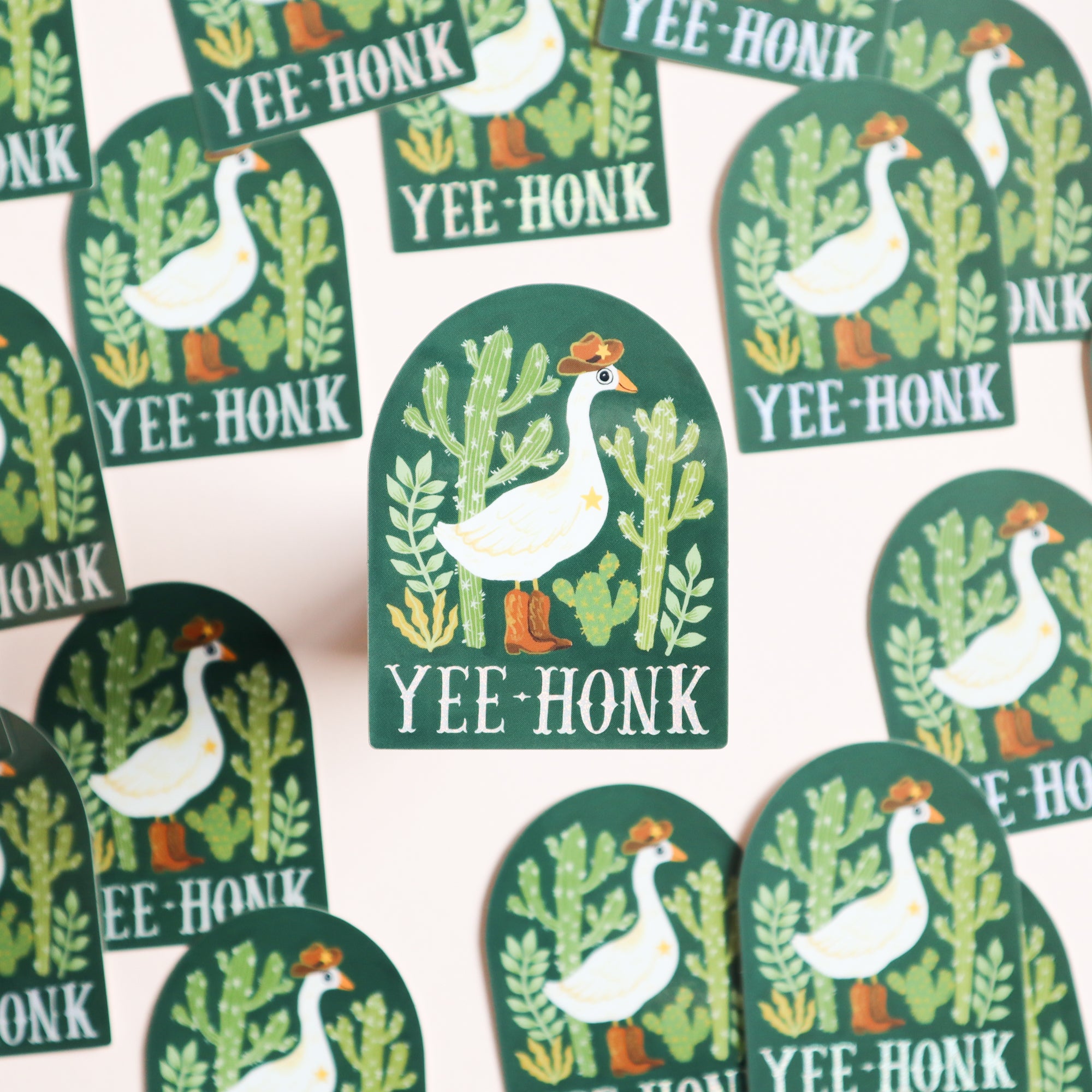 The Yee-Honk Goose Sticker by Papio Press features a cowboy goose in a hat and boots among green cacti with YEE HONK text, set on a light background with a glossy finish for a playful, eye-catching look.