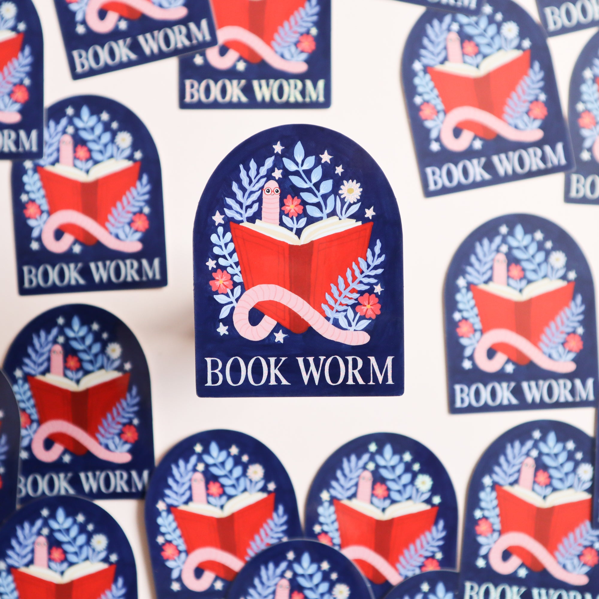 The Papio Press Book Worm Reading Sticker features an open red book with flowers, a cactus, and a pink worm on blue, with BOOK WORM text below. Glossy finish. Perfect for book lovers! Stickers are scattered and overlapping.