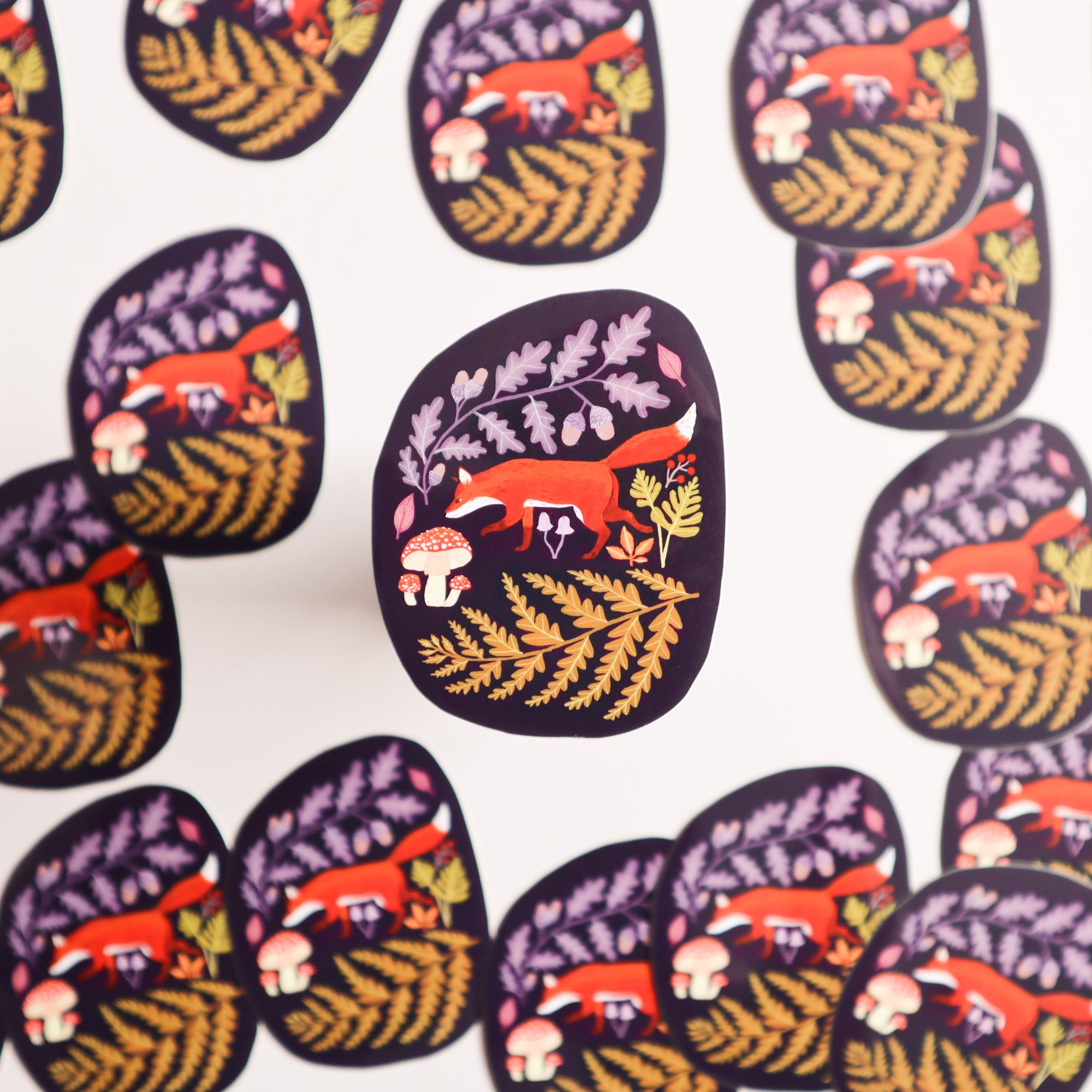 The Autumnal Fox Sticker by Papio Press features an earthy fox, mushrooms, and leaves in a repeating pattern on white vinyl with UV protection. One sticker is shown slightly lifted from the surface.